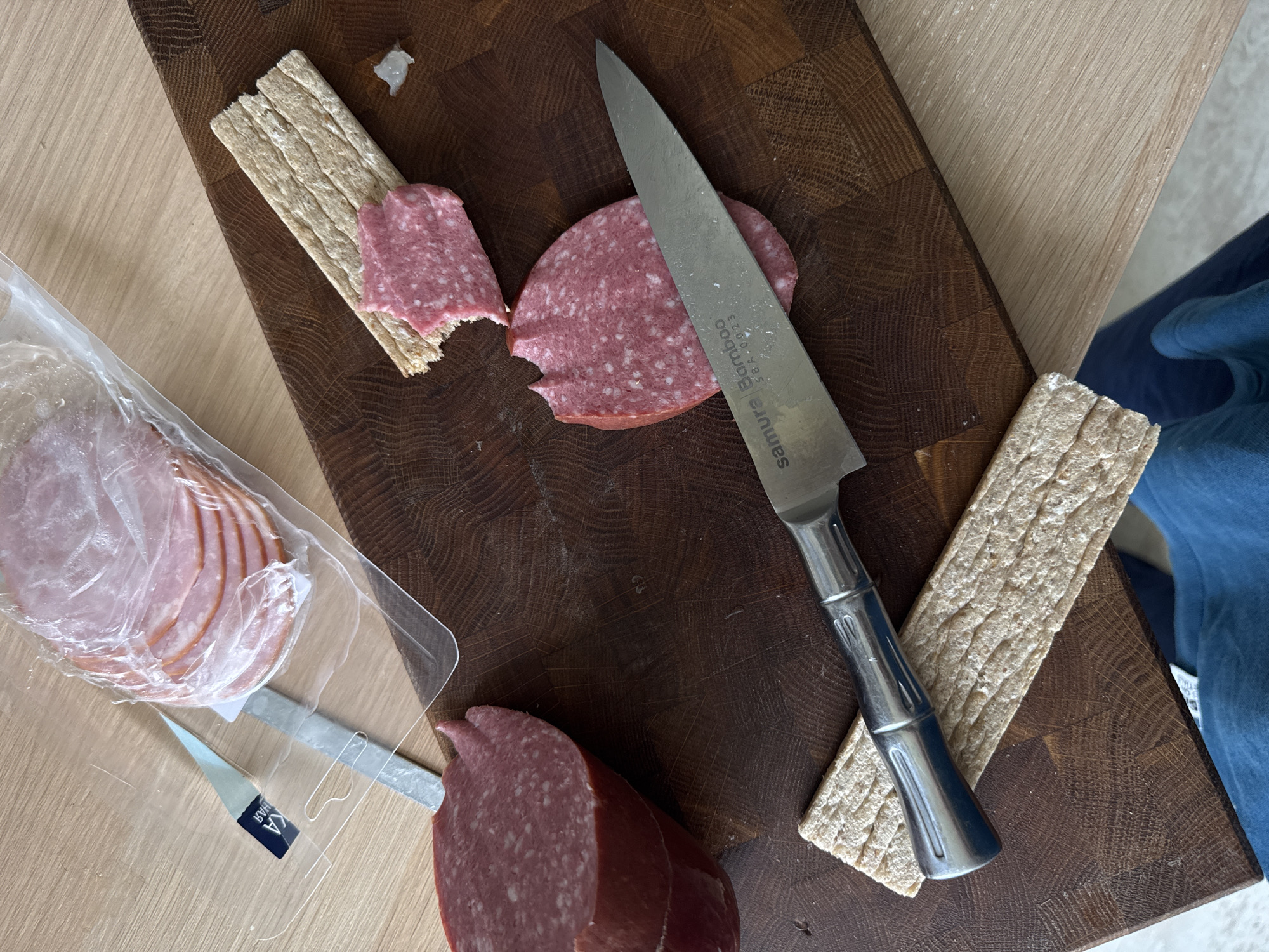This is a top-down, close-up photograph of a wooden cutting board with various food items. The cutting board is made of dark, textured wood and appears to be an end-grain butcher block.
On the cutting board, there are several slices of cured sausage. One large, thick slice is visible on the left, with the cut surface facing upwards. To the right of this, there are two smaller, irregularly shaped pieces of sausage, one of which has a bite taken out of it. A single slice of sausage is positioned beneath a knife.
A silver knife with a bamboo-patterned handle is positioned diagonally across the cutting board, with its blade resting on one of the sausage slices. The text "samura Bamboo SBA-0023" is visible on the blade.
Two pieces of light-colored crispbread are also present on the cutting board. One piece lies horizontally on the lower right, with the knife resting on it. Another piece is positioned above this, partially obscured by the sausage.
In the upper left quadrant of the image, a package of sliced ham is visible, resting on a light-colored wooden surface. The ham slices are thin and pink, and the plastic packaging is slightly crinkled. A small blue and white label with Cyrillic text is attached to the packaging.
The overall impression is a preparation or consumption of a charcuterie-style snack. The image does not contain any identifiable landmarks or features that would place it in Kolomna, Russia.