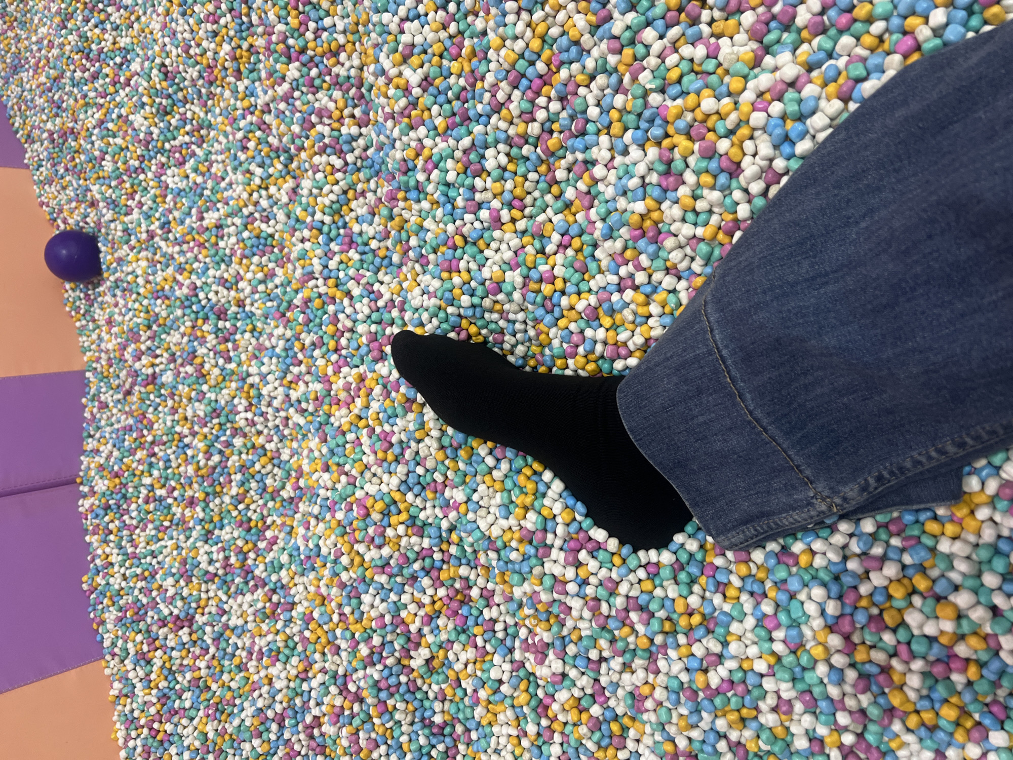 A person's leg, clad in a black sock and blue jeans, rests in a ball pit. The ball pit is filled with small, round, brightly colored balls in shades of pink, blue, yellow, and white. In the background, a purple sphere is visible amongst the balls. Two purple padded walls and a peach-colored wall are also visible in the upper left corner of the image. The location is specified as Kecamatan Pati, Indonesia.