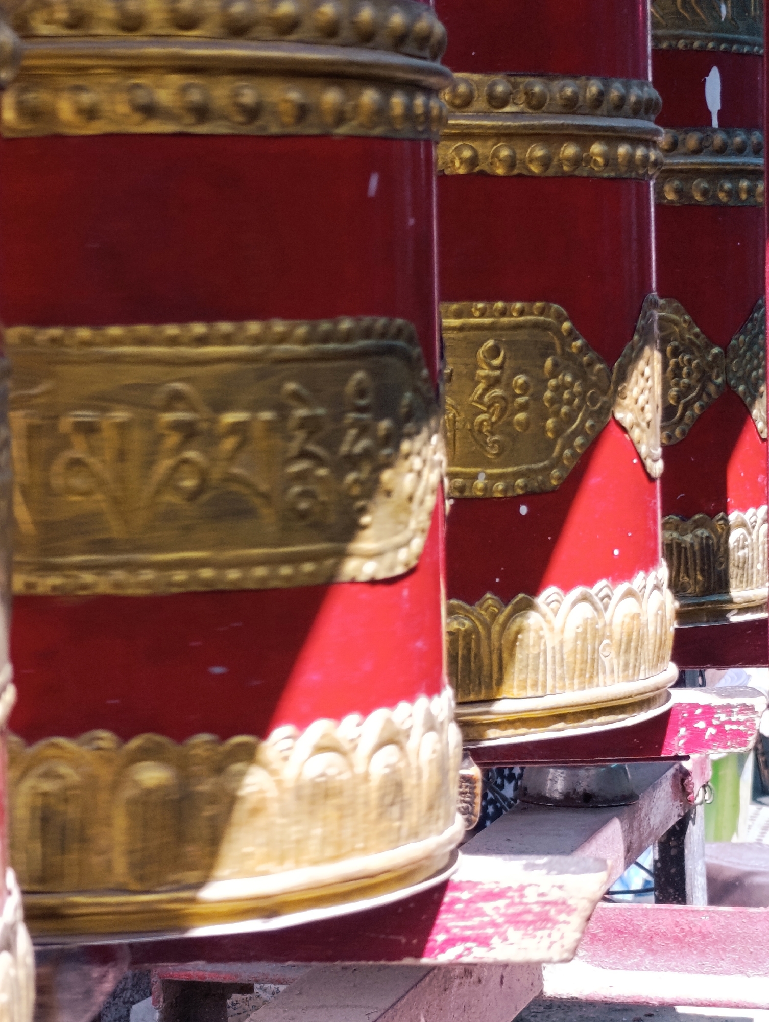 The image shows a close-up of several large, red, cylindrical prayer wheels adorned with intricate gold detailing. The prayer wheels are arranged in a row, suggesting a monastic or religious setting. The gold accents include bands of beads, decorative borders, and embossed script, possibly Tibetan. The surface of the prayer wheels is a vibrant red, and some show signs of wear and white speckling. Sunlight casts shadows across the prayer wheels, highlighting their cylindrical form and the texture of the gold ornamentation. The background is blurred, but suggests a bright, outdoor environment. The prayer wheels are mounted on a wooden structure, also painted red and showing signs of age. The scene is likely located in Leh, India, a city known for its Buddhist monasteries and cultural heritage.