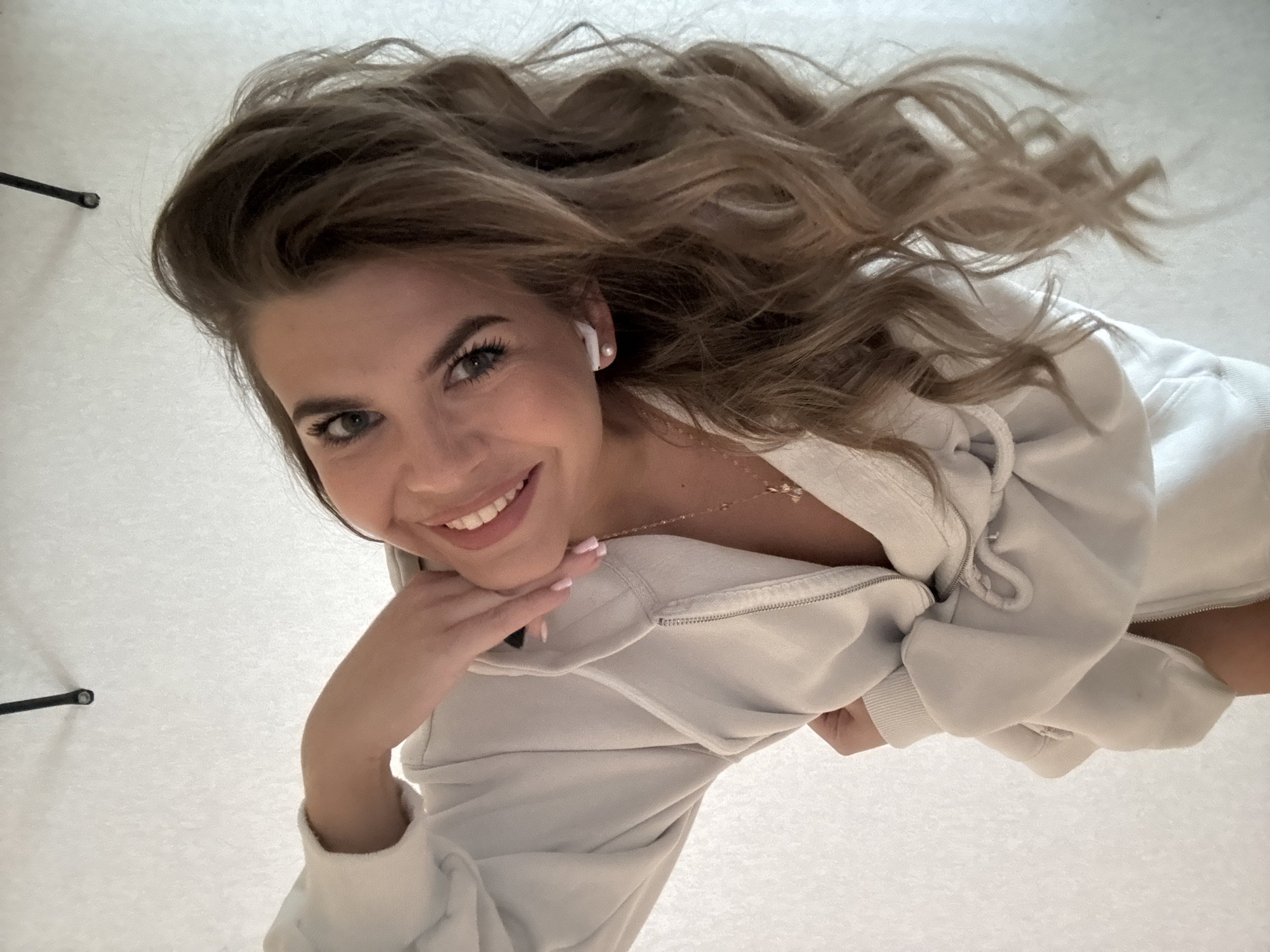 A young woman is captured in a cheerful, low-angle portrait, likely taken from above while she lies down or leans back, set in Chaikovsky, Russia. She is the central subject, looking directly at the camera with a bright, open smile that reveals her white teeth. Her eyes are a light shade, possibly blue or grey.
Her long, voluminous, wavy hair, in shades of light brown or blonde, cascades around her head, filling much of the upper frame. She wears a light-colored (appearing white or very light grey) zip-up hooded sweatshirt, partially unzipped, revealing a delicate gold chain necklace with a small, unidentifiable pendant. A white earbud (AirPod) is visible in her right ear. Her right hand, with neatly manicured nails painted a light pink, is gently placed under her chin.
The background is a plain, uniform, and light-colored surface, possibly a bed, floor, or carpet, which keeps the focus entirely on the woman. Faint dark shapes, resembling parts of a stand or furniture leg, are visible at the extreme top and bottom left edges of the frame. The lighting is soft and even, creating a natural and inviting atmosphere. The overall emotion conveyed is happiness and relaxation. There is no visible text in the image.