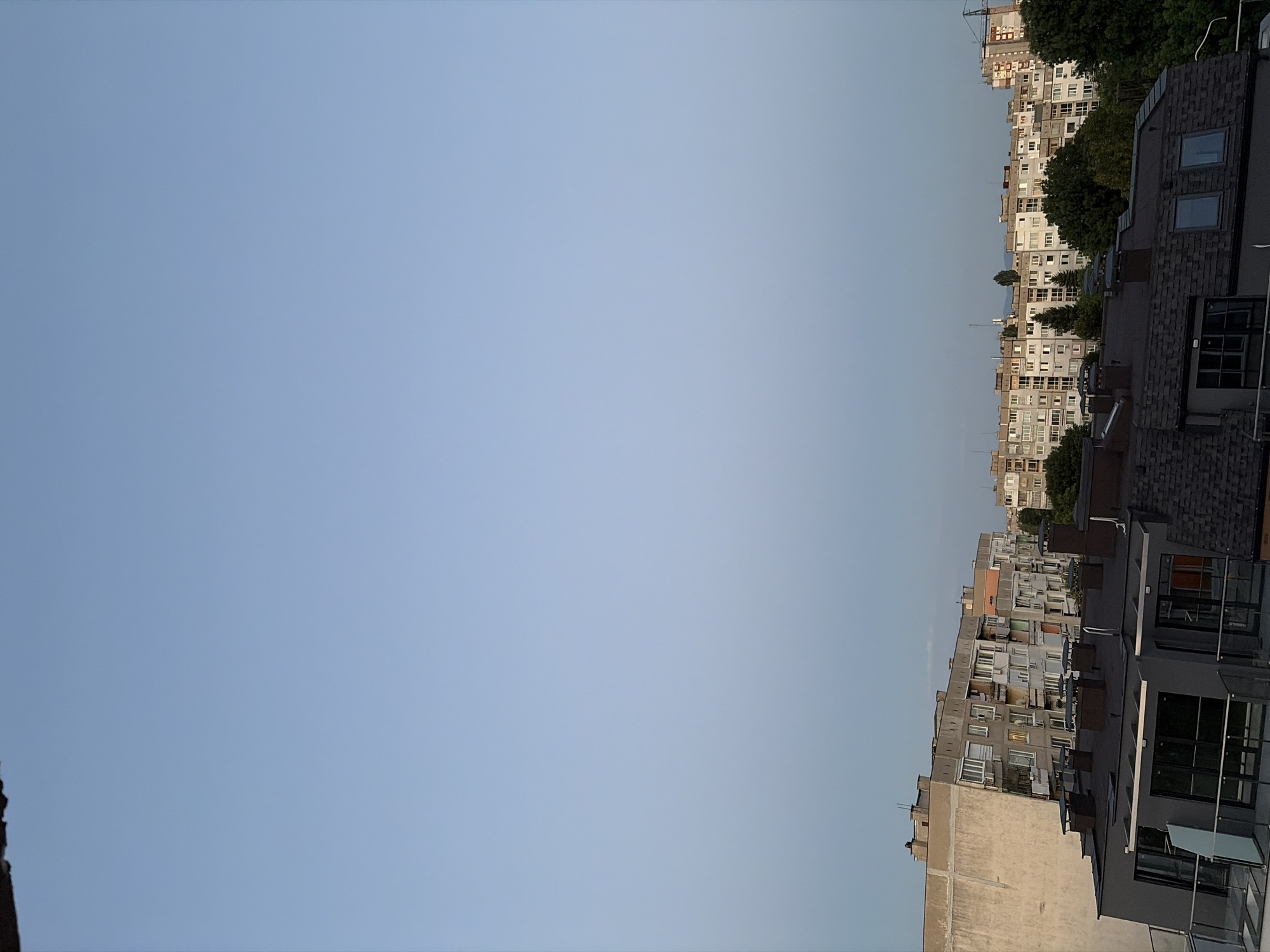 An eye-level, vertical outdoor shot captures an urban skyline under a pale blue sky. The sky occupies the majority of the frame, with the top edge showing a sliver of a dark roof overhang. The lower portion of the image displays a dense collection of residential buildings of varying architectural styles and ages. In the foreground, a dark gray building with a modern design features large glass windows and a dark, pitched roof with numerous square, dark brown chimney pots. Behind it, a series of older, block-style apartment buildings dominate the mid-ground. These buildings are characterized by their uniform facades, multiple windows, and balconies, with some showing signs of wear and tear. To the left, a tall, lighter-colored building with a weathered facade stands out. A line of lush green trees forms a natural boundary between the apartment blocks and the sky. Further in the distance, more buildings and a construction crane are visible. The lighting suggests it is late afternoon or early evening, with a soft, diffused light illuminating the scene. There are no people or animals visible in the frame.