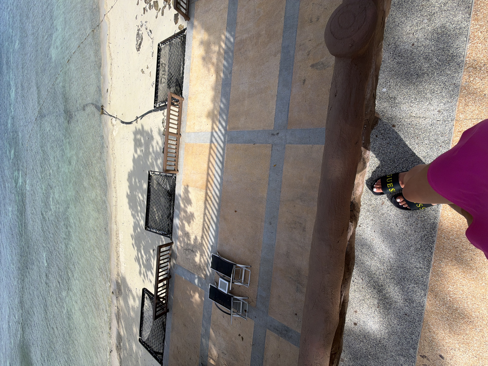 The image shows a view from an elevated position looking down at a beach and a patio area in Ao Nang, Thailand. The beach is sandy with clear turquoise water. There are buoys marking an area in the water. On the sandy beach, there are several black hammock-like chairs with wooden frames. Adjacent to the beach is a paved patio area with a tiled surface. On the patio, two black lounge chairs with metal frames are set up, along with a small side table. In the foreground, a person's legs are visible, wearing black "Guess" slides and a bright pink garment, standing on a stone pathway. The patio is partially covered in shadows cast by an unseen structure.