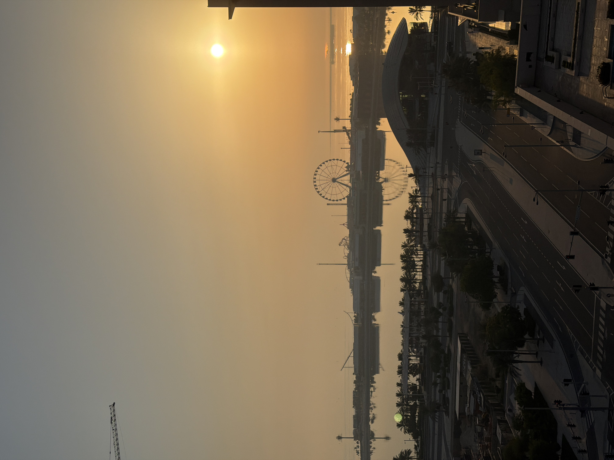 The image shows a city view at sunrise. The sun is a bright orb in the sky, casting a warm orange glow over the scene. In the distance, a Ferris wheel and other amusement park rides are visible, along with buildings and palm trees. The water in the foreground reflects the sky and the structures. A road with streetlights and traffic signals is in the lower part of the image, with buildings and construction sites on either side. A crane is visible in the upper left corner, suggesting ongoing construction in the area. The overall impression is of a modern city waking up to a new day.
