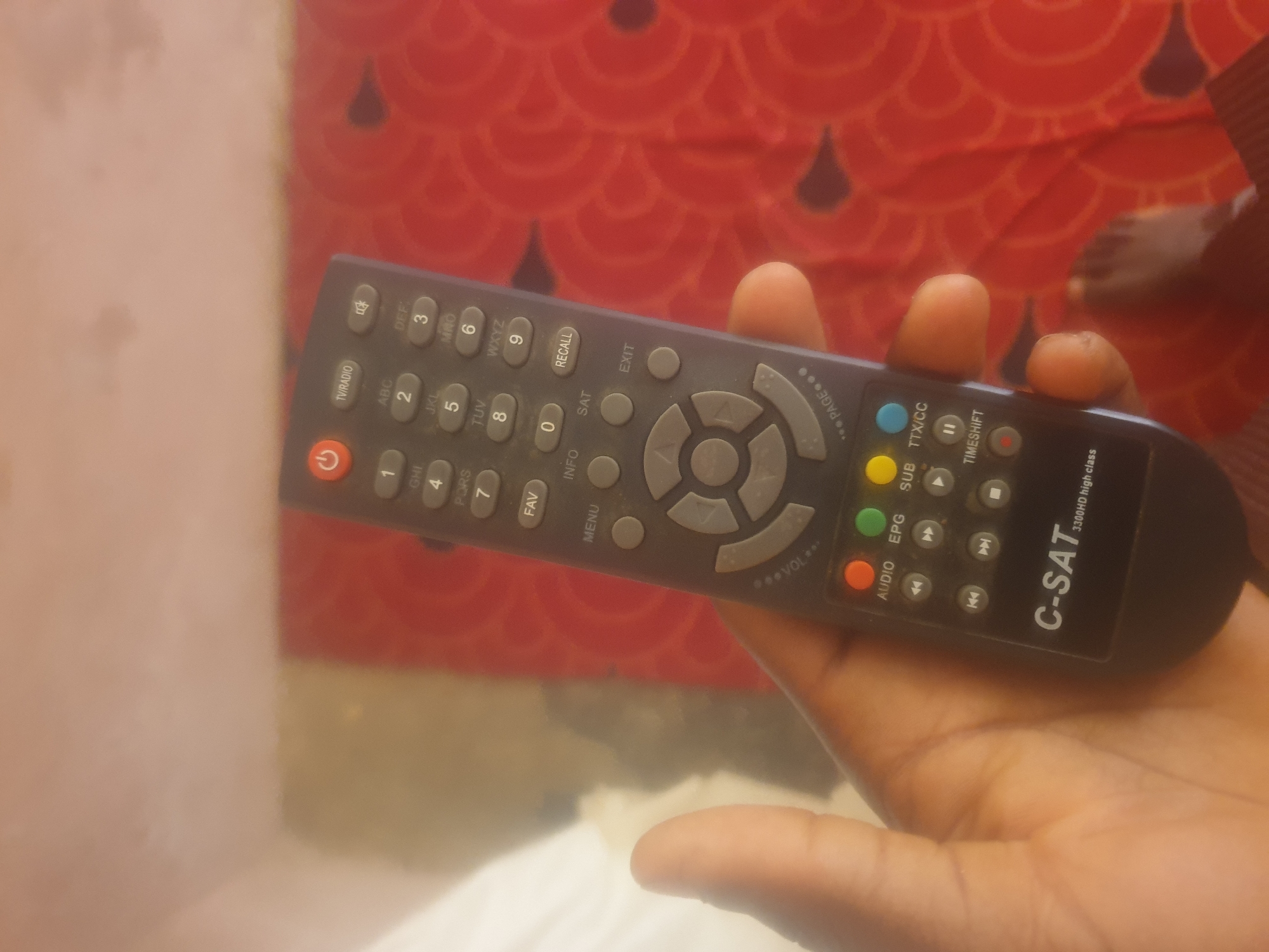 A hand holds a black remote control with various buttons and labels in front of a red patterned cloth. The remote has a power button on top, followed by a numeric keypad with labels like "TV/RADIO," "ABC," "DEF," "GHI," "JKL," "MNO," "PORS," "TUV," and "WXYZ." Below the numbers are buttons labeled "FAV," "INFO," "RECALL," "MENU," "SAT," and "EXIT." A circular navigation pad with directional arrows is present in the middle. Towards the bottom, there are colored buttons (red, green, yellow, blue) labeled "AUDIO," "EPG," "SUB," and "TTX/CC." Other buttons include rewind, play/pause, fast forward, stop, and a "TIMESHIFT" button. The brand "C-SAT 3300HD high class" is printed at the bottom of the remote. The image does not provide any information to determine its location in Washington, United States of America, or any other specific city.
