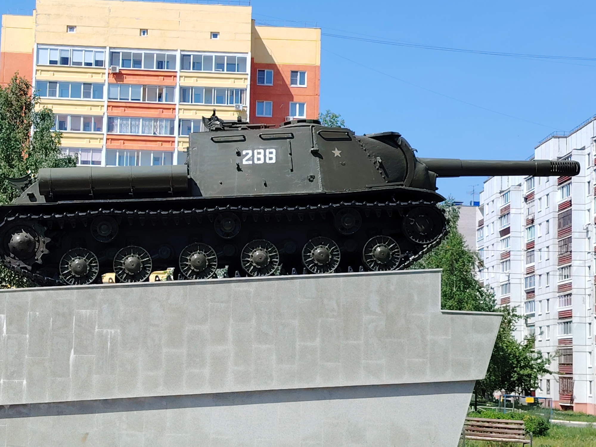 The image depicts a self-propelled gun, specifically a Soviet ISU-152 tank destroyer, serving as a monument in Chaikovsky, Russia. The vehicle, painted in dark green, is mounted on a concrete pedestal. The hull displays the number "288" in white. The gun barrel is extended forward. Behind the tank are residential buildings with numerous windows. The sky is clear and blue. The surrounding area includes trees, grass, and a park bench.