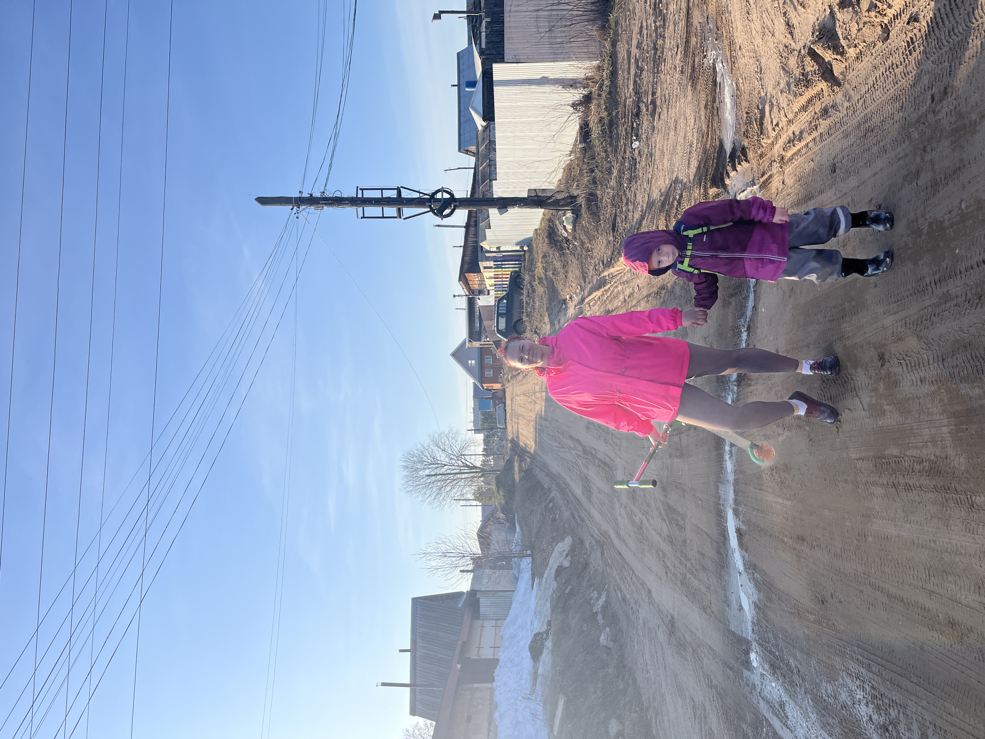 Two people stand on a dirt road in Chaikovsky, Russia. An adult in a bright pink jacket holds the hand of a child in a purple hooded jacket. The road is muddy, with tire tracks and patches of melting snow. A woman is using a scooter. Utility poles line the road, and houses are visible in the background. The sky is bright blue.