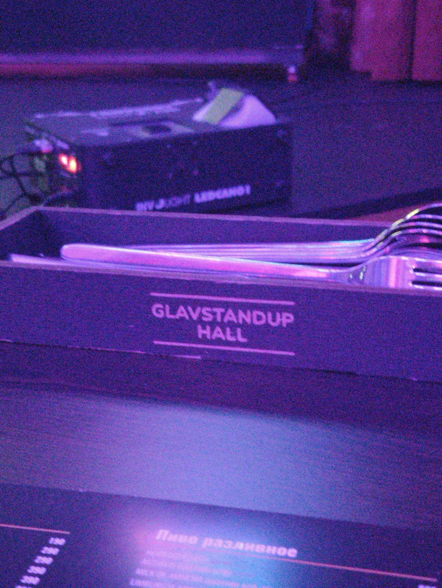 The image depicts a close-up view inside "Glavstandup Hall", a stand-up comedy venue located in Moscow, Russia. Forks and cutlery are resting on the counter. A menu board showing the price of beer is also partially visible.