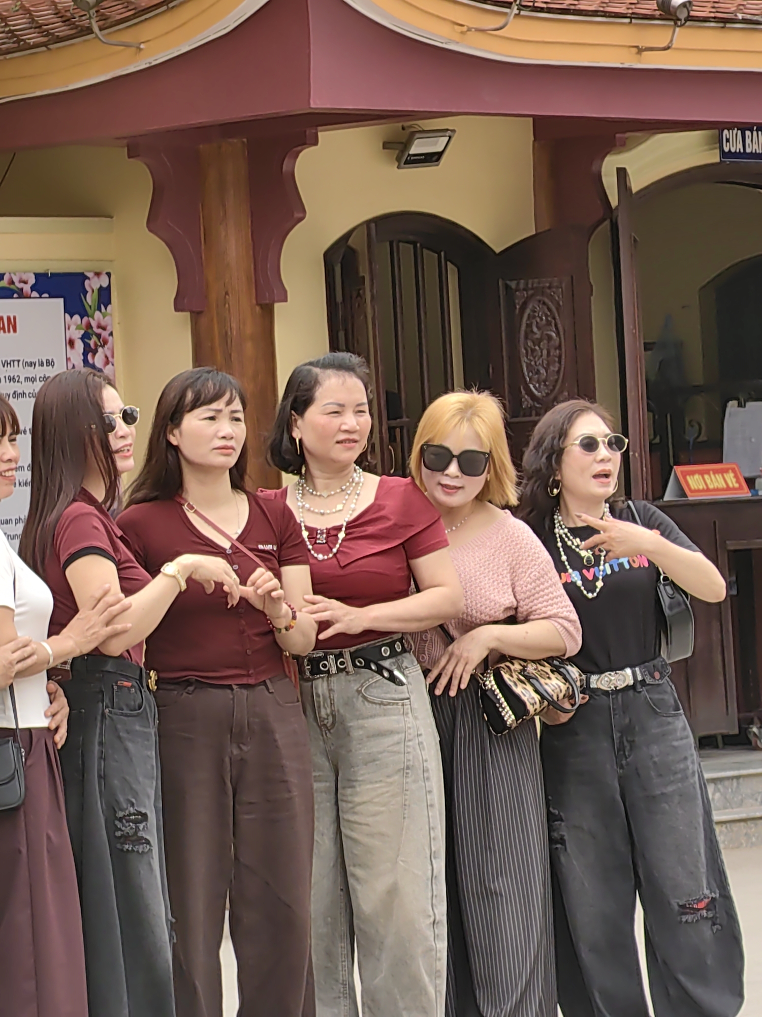 Six women are posing together in front of a building. The building has a sign that reads "CỬA BẢN". Another sign is visible that includes the text "VHTT". The women are wearing various outfits including maroon tops, a pink sweater, a black T-shirt, and jeans. Some are wearing sunglasses and necklaces. One woman is carrying a leopard print purse. The building has an ornate wooden door and column details. The scene appears to be in Lạng Sơn, Vietnam.