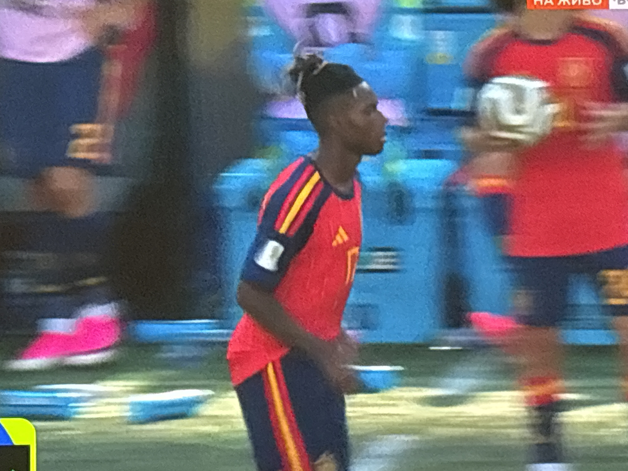 The image captures a portion of a soccer match. A young Black male athlete, wearing a red and blue soccer jersey with yellow accents, is in the foreground, turned to his right and looking off-camera. His hair is styled in a bun atop his head. He is wearing dark blue shorts with a yellow and red stripe down the side. His left foot, clad in a bright pink soccer shoe, is visible near the bottom of the frame. In the background, another player in a red jersey is holding a white soccer ball. The environment appears to be a soccer field with a blurred blue backdrop, possibly representing stadium seating or screens. The overall impression is one of motion and athletic activity. The image does not contain any recognizable landmarks or indications of the city of Burgas, Bulgaria.