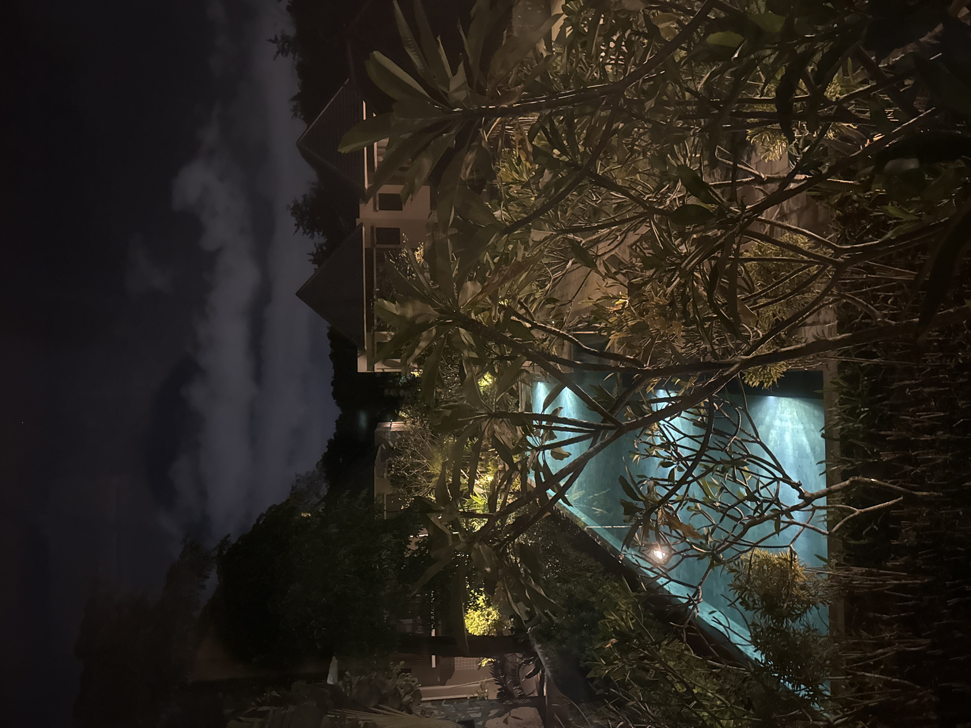 At night, the image depicts a swimming pool located in Kuta Selatan, Indonesia. The turquoise-colored water in the pool is illuminated, likely by underwater lights. Overhanging the pool are branches of trees and foliage, partially obscuring the view. In the background, a building with multiple stories and a sharply angled roof can be seen. The sky is dark, with some visible clouds.