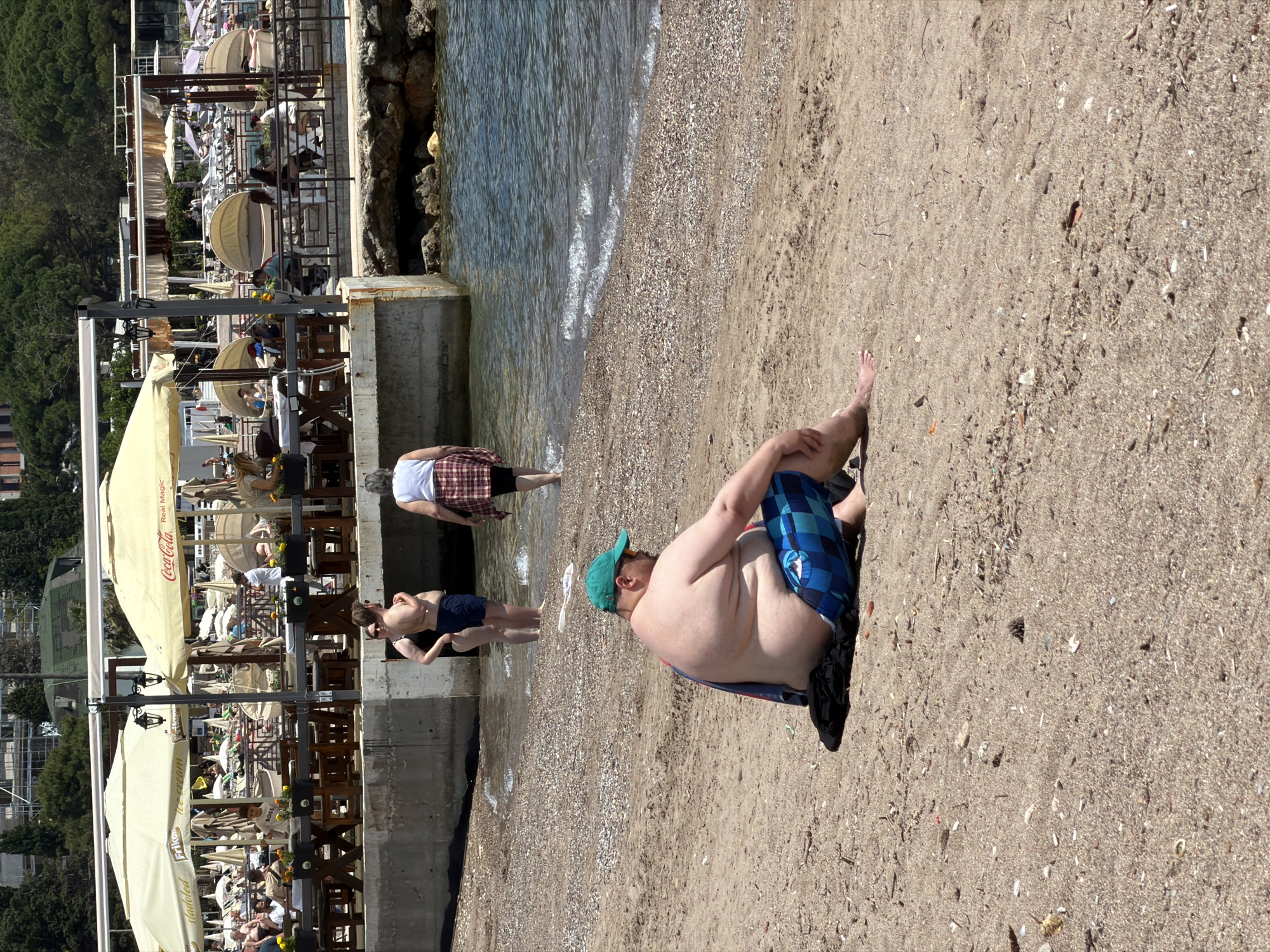 The image depicts a beach scene in Budva, Montenegro. Three people are standing in the shallow water by a concrete wall. There is a man sitting on the sand wearing blue checkered shorts and a green cap. A black bag is beside him on the sand. Further down the beach, multiple rows of lounge chairs and umbrellas are set up for tourists. There is an area with wooden tables and chairs. Buildings and trees are visible in the background.