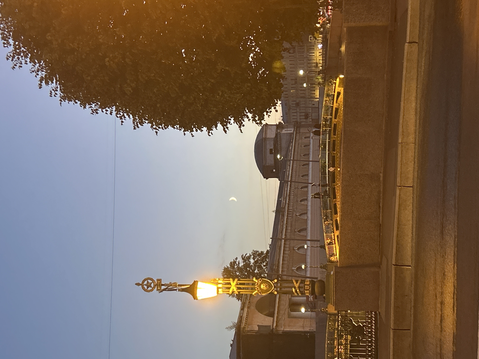 A lamppost with an ornate gold finial featuring a star and a crescent moon stands illuminated against a clear, pale blue sky at dusk. The lamppost's lantern emits a warm yellow light. In the background, a long, tan building with arched windows runs horizontally, and a domed structure is visible to its left. A crescent moon hangs in the sky. A bridge with illuminated railings spans a body of water in the mid-ground, with people visible on it. To the right, a large tree with dark green leaves partially obscures the view. The foreground shows a street or walkway with a textured surface. The overall scene depicts an urban setting transitioning from daylight to evening.