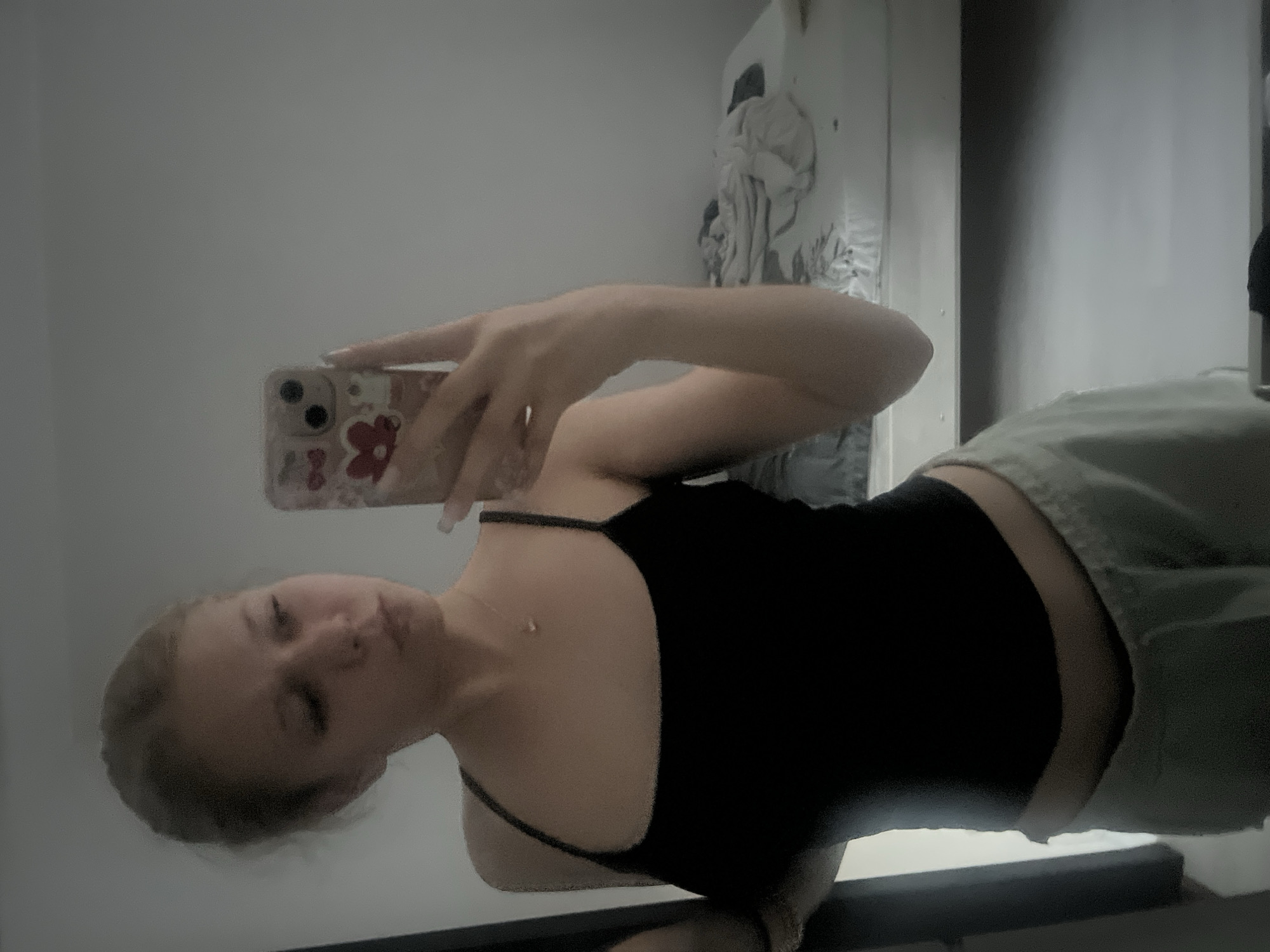 A young blonde woman is taking a mirror selfie in a room. She is wearing a black spaghetti strap top and light grey sweatpants. Her hair is pulled back, and she is wearing a delicate necklace. She is holding a cell phone with a patterned case. In the background, a bed with rumpled sheets is visible. The room appears to be a bedroom or living space.