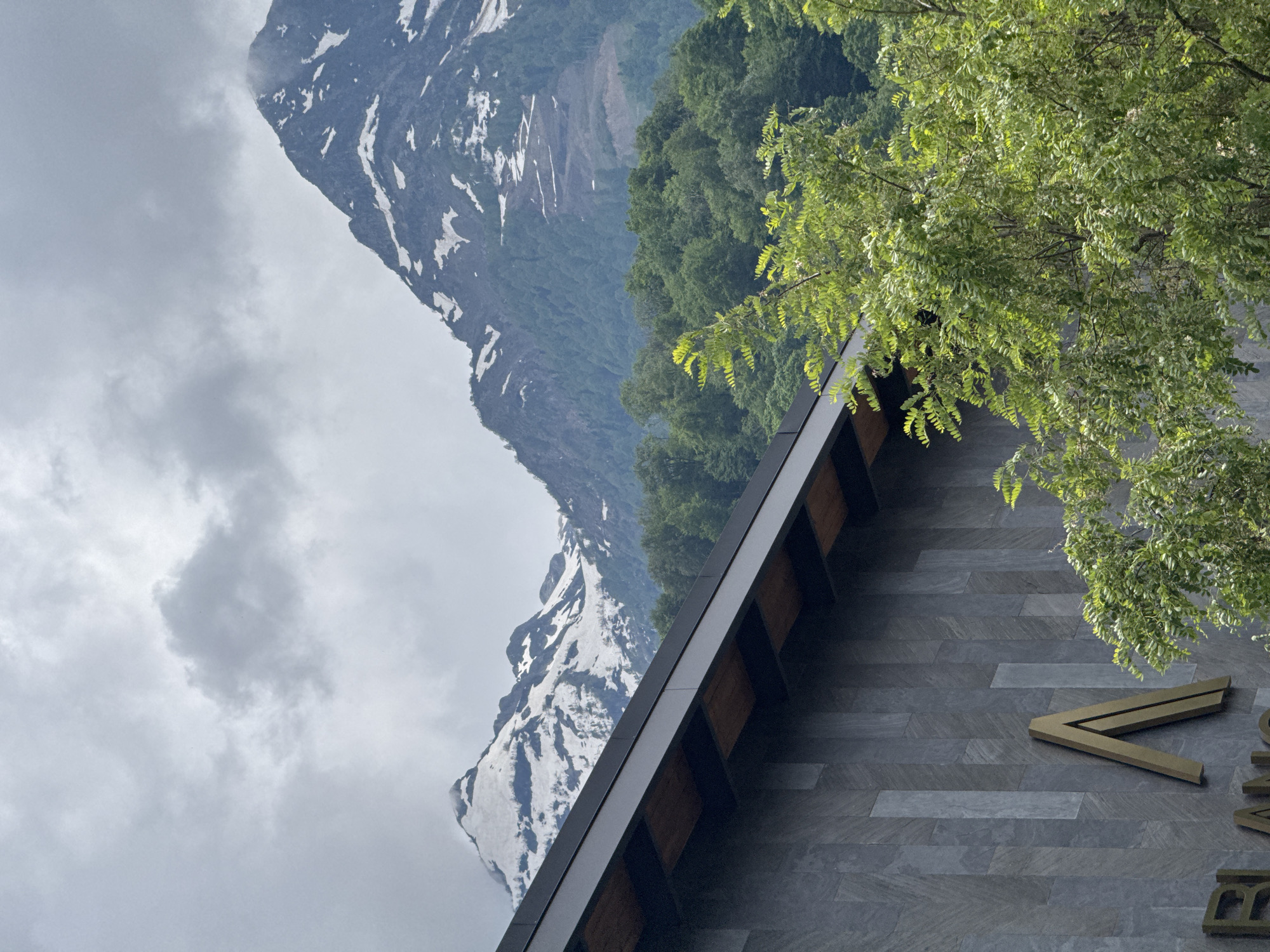 The image shows a building with a dark gray, textured facade, featuring vertical panels and a black roof with wooden accents. A prominent golden "A" logo is mounted on the facade, along with partial lettering that appears to read "BLAN." A lush green tree with light green leaves is in the foreground, partially obscuring the building. In the background, a majestic mountain range with snow-capped peaks rises against a cloudy sky. The overall scene suggests a location in a mountainous region, likely a resort area, with the building potentially being a hotel or commercial establishment. The location is identified as Estosadok, Russia.