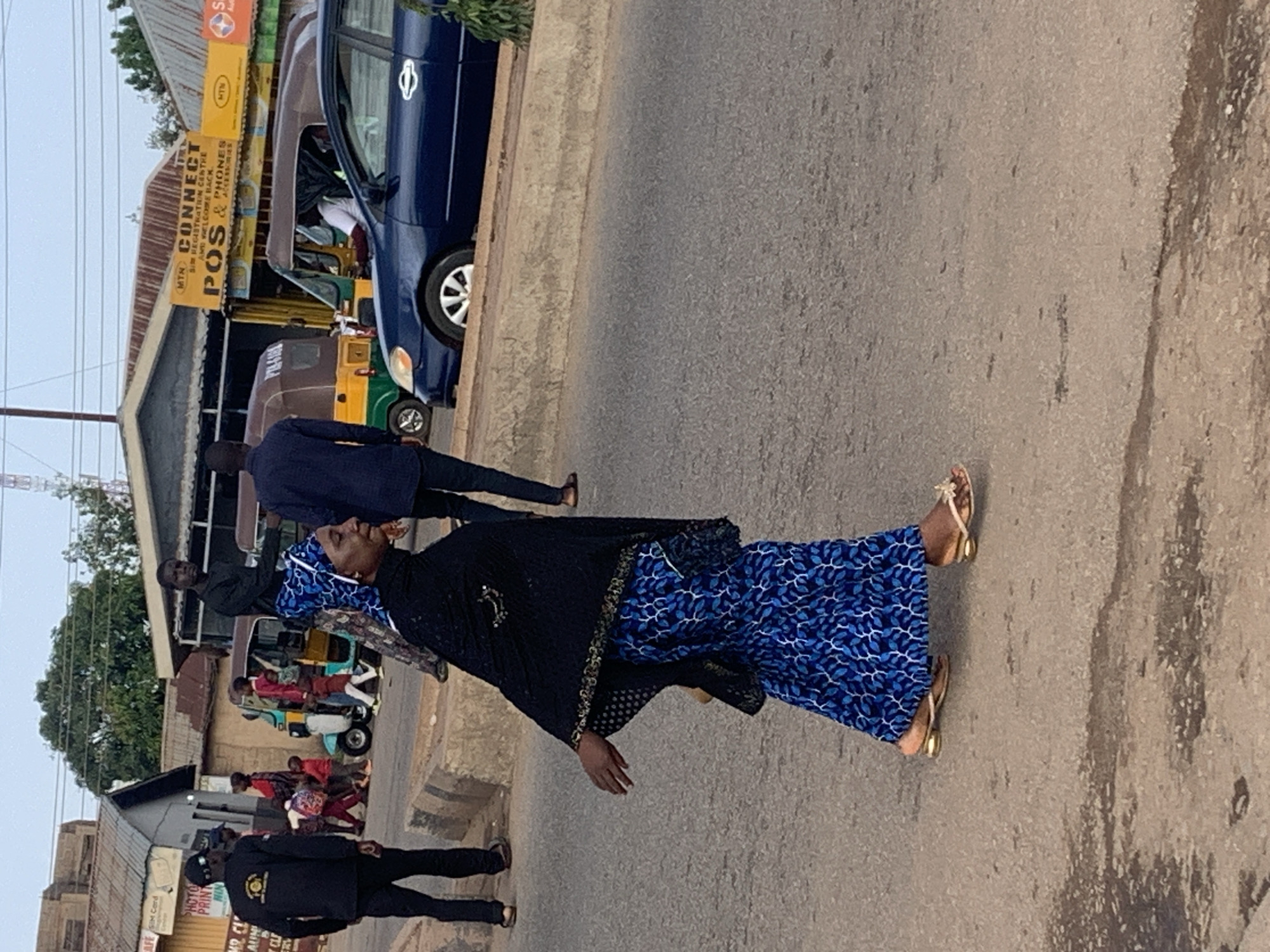 In the city of Gura Top, Nigeria, an outdoor scene unfolds on a paved road during daylight hours. A woman, dressed in a blue patterned dress and a black headscarf with gold trim, walks across the road. She wears gold-heeled sandals. To her right, a dark blue sedan is parked on the other side of a low concrete barrier. In the background, several people are visible, including a man in a black shirt and dark pants walking towards the left and two other individuals further in the distance. A yellow and green tuk-tuk is also present. Buildings with corrugated metal roofs line the street, and a yellow sign with the text "MTN CONNECT POS & PHONES" is affixed to one of them. The sky is overcast.