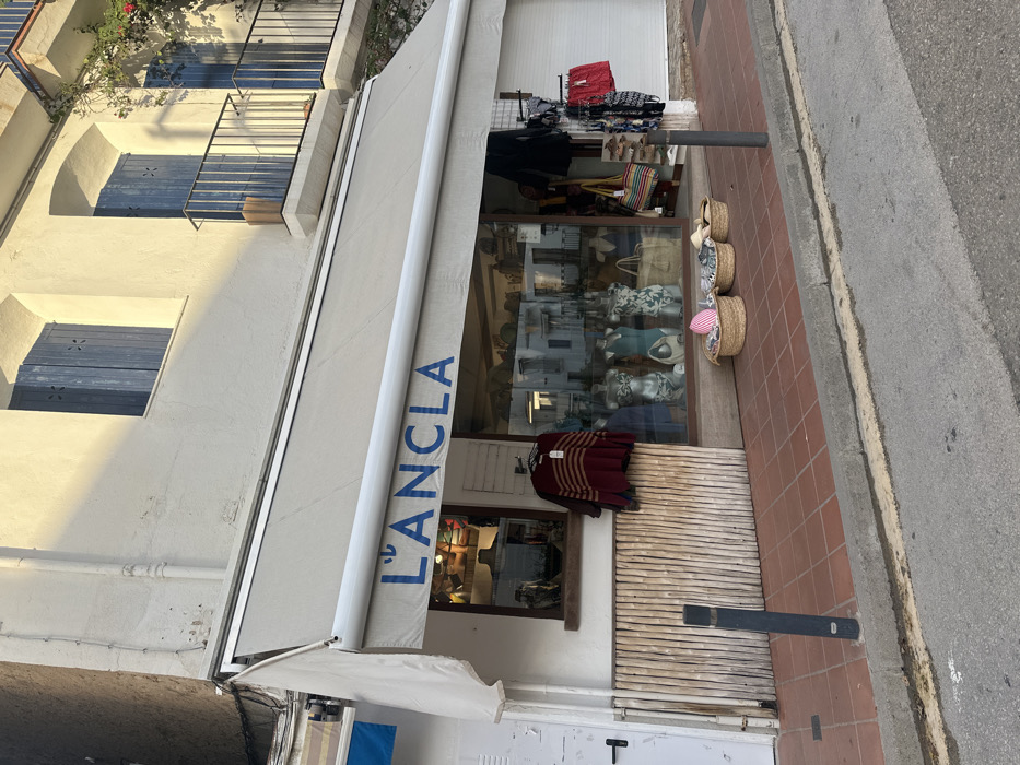 The image depicts the storefront of a shop named "L'ANCLA" located in Calella De Palafrugell, Spain. The building features a white stucco facade with several windows, including two on the upper level with closed blue wooden louvered shutters and a third on the right with a small black iron balcony. A second-floor balcony with a similar railing is visible further right, partially obscured by green foliage with red flowers.
The ground floor storefront is covered by a retractable beige awning. Below the awning, the shop's name, "L'ANCLA," is prominently displayed in blue capital letters. The shop window displays several items, including three headless mannequins wearing patterned swimwear and various bags and accessories. Inside the shop, reflections from the opposite side of the street are visible in the glass.
To the left of the main display window, a smaller window is present, with a male torso mannequin visible inside. Below this window, the lower portion of the wall is clad with vertical light-colored wooden or bamboo slats. A dark-colored garment with horizontal red and beige stripes hangs on a hook outside the shop, to the left of the main display.
To the right of the main shop window, a black metal grid rack holds various clothing items, including patterned tops and red shorts. Directly in front of the shop window, on the tiled terracotta sidewalk, three circular woven baskets contain merchandise such as striped cushions and hats.
The sidewalk is paved with terracotta-colored tiles, leading to a grey concrete curb. A black and grey bollard is fixed into the sidewalk near the left side of the frame. Beyond the curb, an asphalt road with visible wear and light colored markings is present. The scene is illuminated by strong sunlight from the right, casting shadows on the building and highlighting textures. No individuals are visible in the frame.