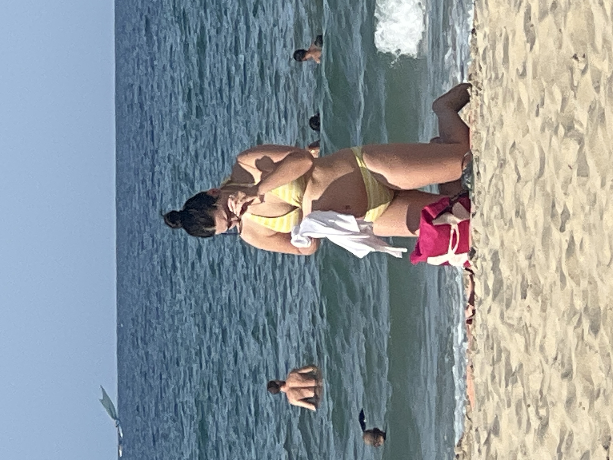A woman in a yellow striped bikini is kneeling on a sandy beach, holding a white garment. She is facing down, and her hand is near her face, possibly wiping her eyes or nose. To her left, a pink backpack is visible on the sand. The ocean stretches behind her, with several people swimming in the water. In the distance, a sail or kite-like object is visible on the horizon, and a wave is breaking on the right side of the frame. The beach appears to be in Da Nang, Vietnam, based on the context provided.