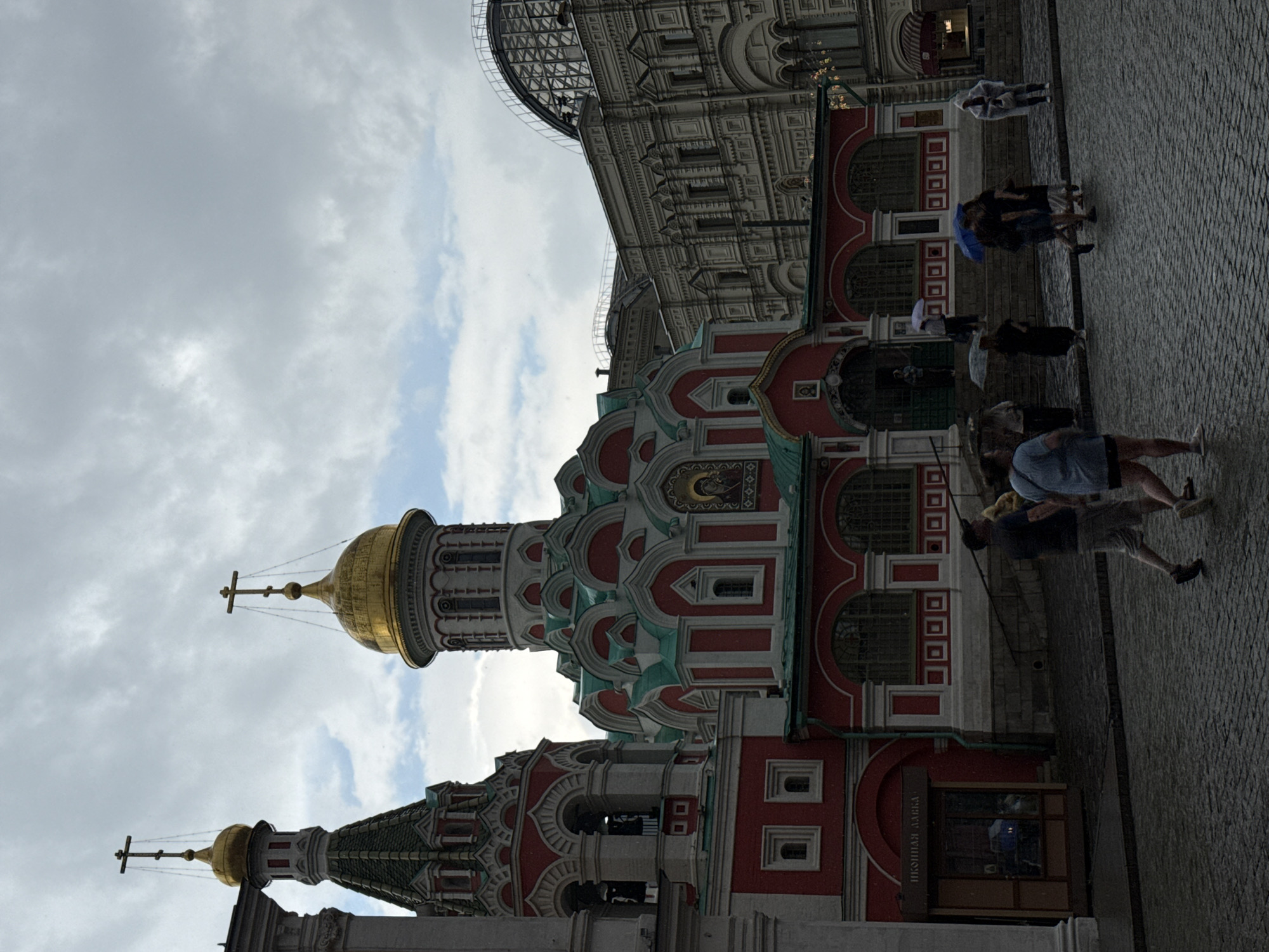 The image depicts an overcast day in Moscow, Russia, with a view of the iconic Saint Basil's Cathedral on Red Square. The cathedral is characterized by its vibrant red and white facade, intricate architectural details, and a collection of colorful onion domes topped with golden crosses. To the left of the cathedral, a portion of the Moscow Kremlin wall is visible, featuring battlements and towers. In the foreground, cobblestone streets are wet from rain, with several pedestrians holding umbrellas and walking in different directions. The people are dressed in casual attire, and some are captured mid-stride. The overall atmosphere is somber due to the weather, yet the architectural grandeur of the cathedral remains the focal point.
