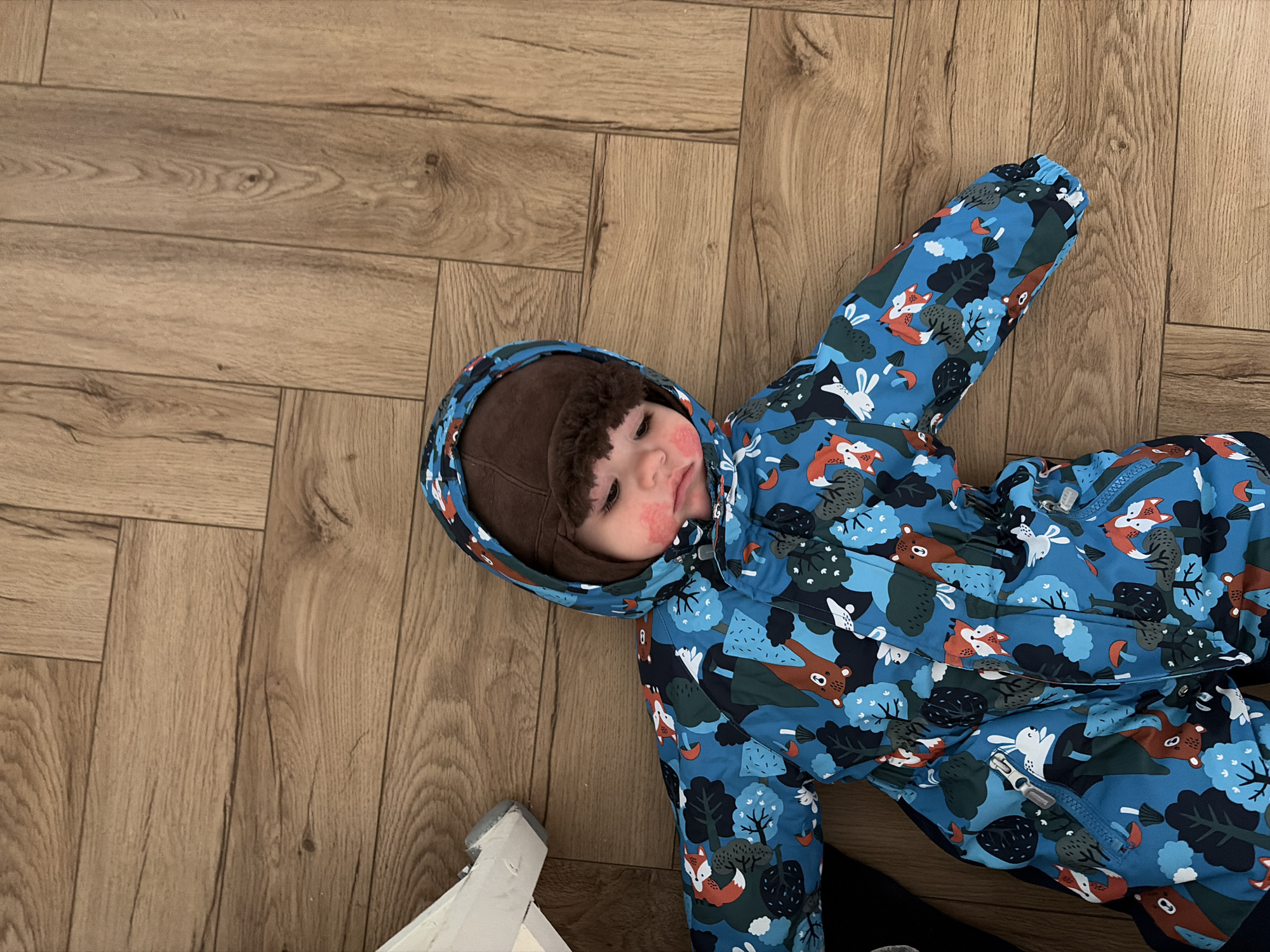 A toddler in a blue patterned snowsuit with a brown hat lies on a wood-patterned floor in an indoor setting. The snowsuit has cartoon-like forest animal and tree motifs. Part of a white object is visible in the lower-left corner. The toddler has rosy cheeks. Location: Chaikovsky, Russia.