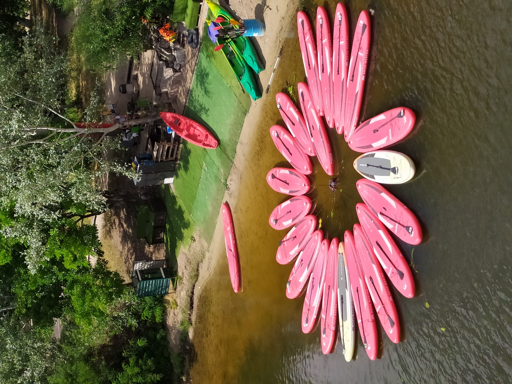 In Kyiv, Ukraine, a scenic outdoor recreation area on the Dnipro River features numerous pink stand-up paddleboards (SUPs) arranged in a circular pattern in the water, some with paddles. Several green kayaks are on the sandy bank, along with a red kayak. Beyond the water, a grassy area transitions to a sandy embankment with trees, a small structure, and some seating. The scene appears to be a watersports rental or activity center.