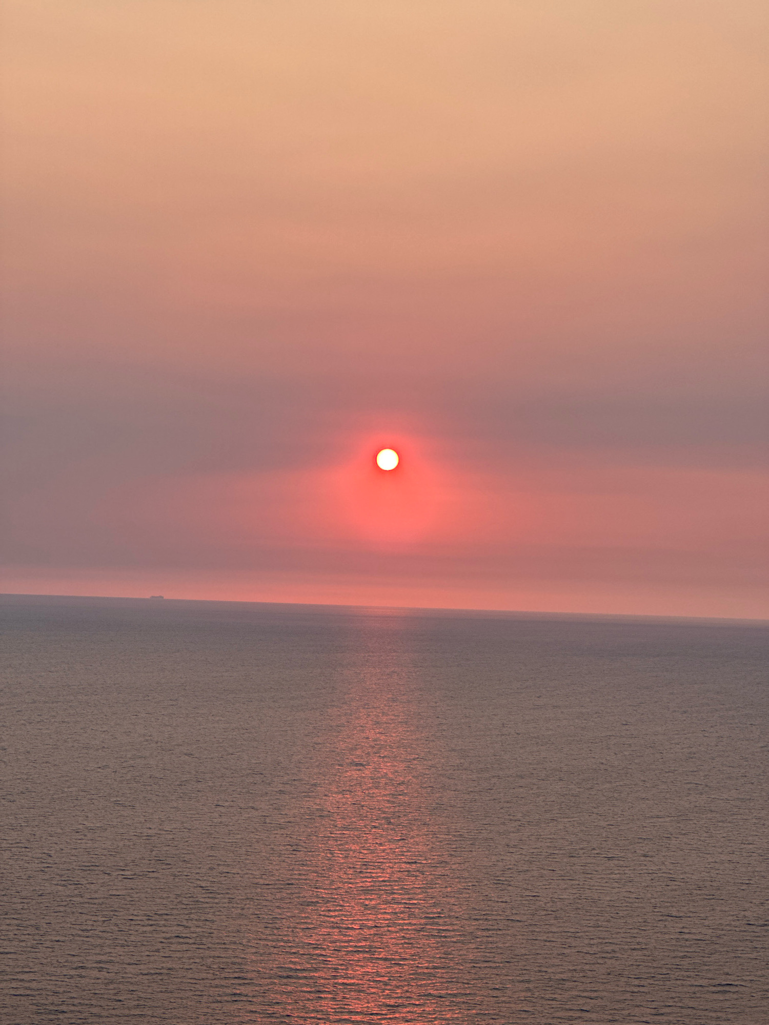 A solitary sun, a bright orange orb, hangs low in a hazy pink sky, casting a shimmering reflection on the calm, gray sea. The horizon is a faint line, with a distant, dark silhouette of a ship visible on the left. The overall impression is one of serene, muted twilight. The scene is not located in Zagora, Montenegro.