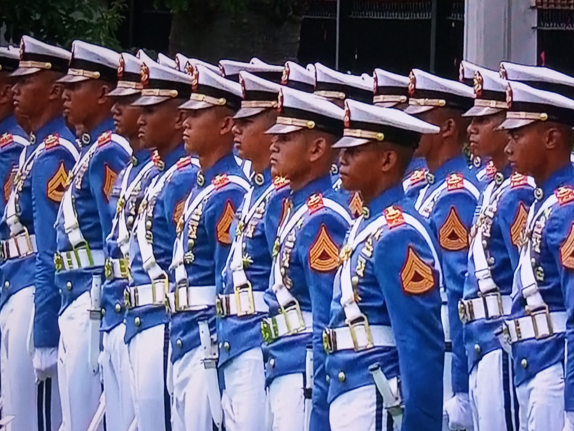 The image shows a formation of soldiers in uniform standing at attention. They are wearing blue jackets with white sashes, white belts with gold buckles, and white trousers. Each soldier also wears a white hat with a gold band. Some soldiers have a dark colored band on the cuff of their sleeves and a gold insignia on their upper arm. Several soldiers have a dagger or bayonet tucked into their belt. The soldiers are standing in a line, facing forward, with their heads turned slightly to their left. The background appears to be an outdoor setting with trees and buildings visible. The image is a medium shot, capturing the soldiers from the waist up. The scene suggests a formal military event or parade, possibly in the city of Banyudono, Indonesia.