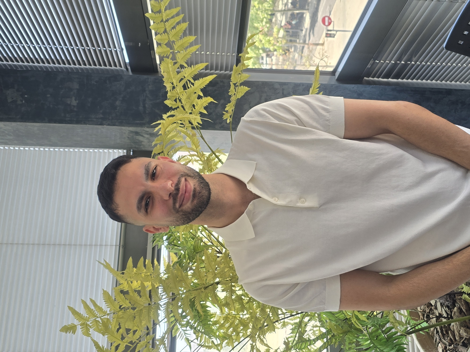 A medium shot captures a man from the chest up, standing indoors with a potted fern to his left and blinds to his right. He has dark hair and a beard, and is smiling at the camera. He is wearing a white polo shirt. Behind him, through a window, is a cityscape with trees, buildings, and a street with a red traffic signal. The man is positioned in front of a dark grey pillar. The overall lighting suggests an indoor setting during the day. The location is identified as Terrassa, Spain.
