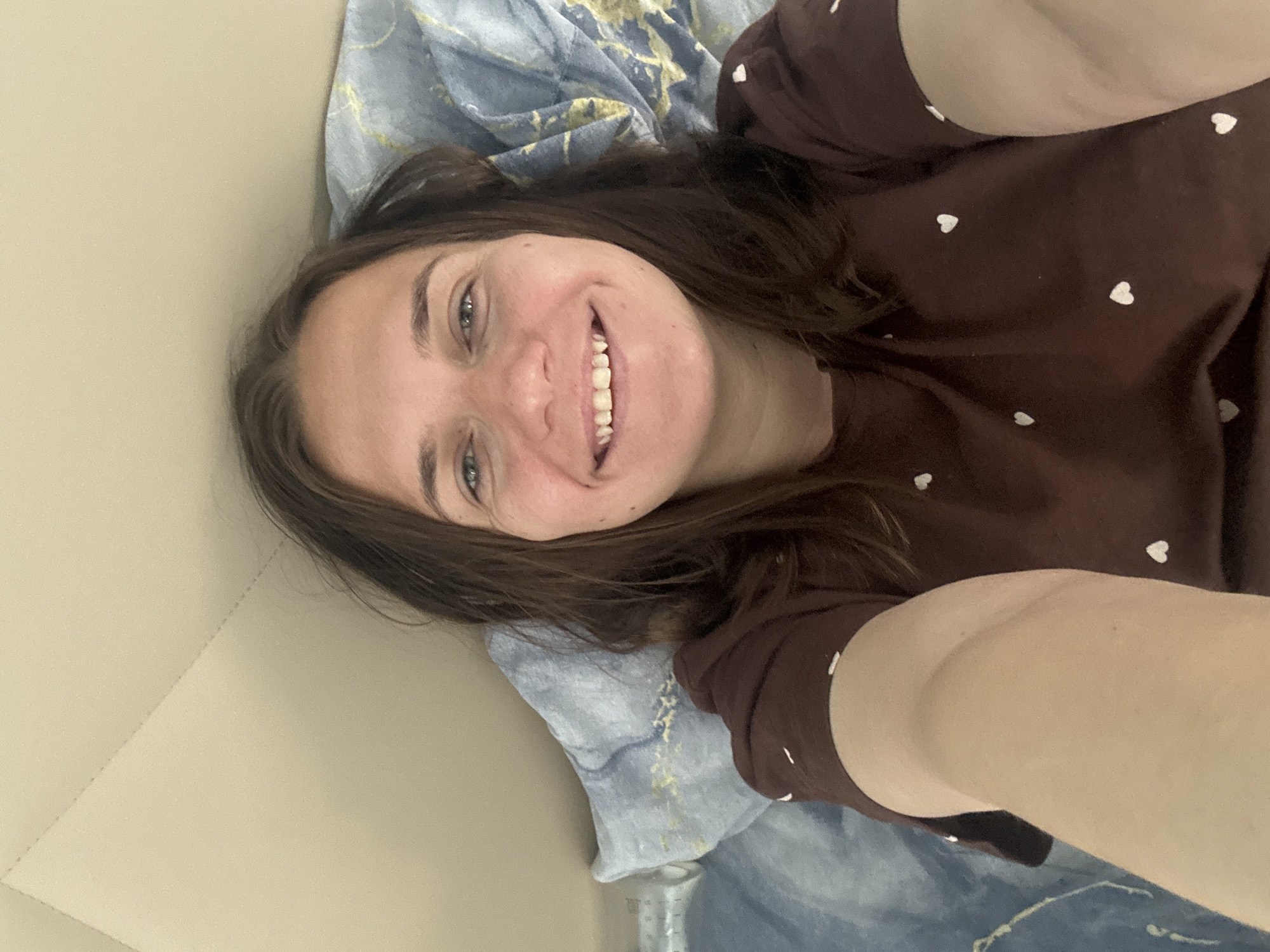 A selfie taken by a woman with long brown hair, lying on her back on a patterned blue and white surface. She wears a brown top with small white hearts. Her face is turned towards the camera, and she is smiling, revealing her teeth. The location, Раздолье, Russia, is not discernible from the image.