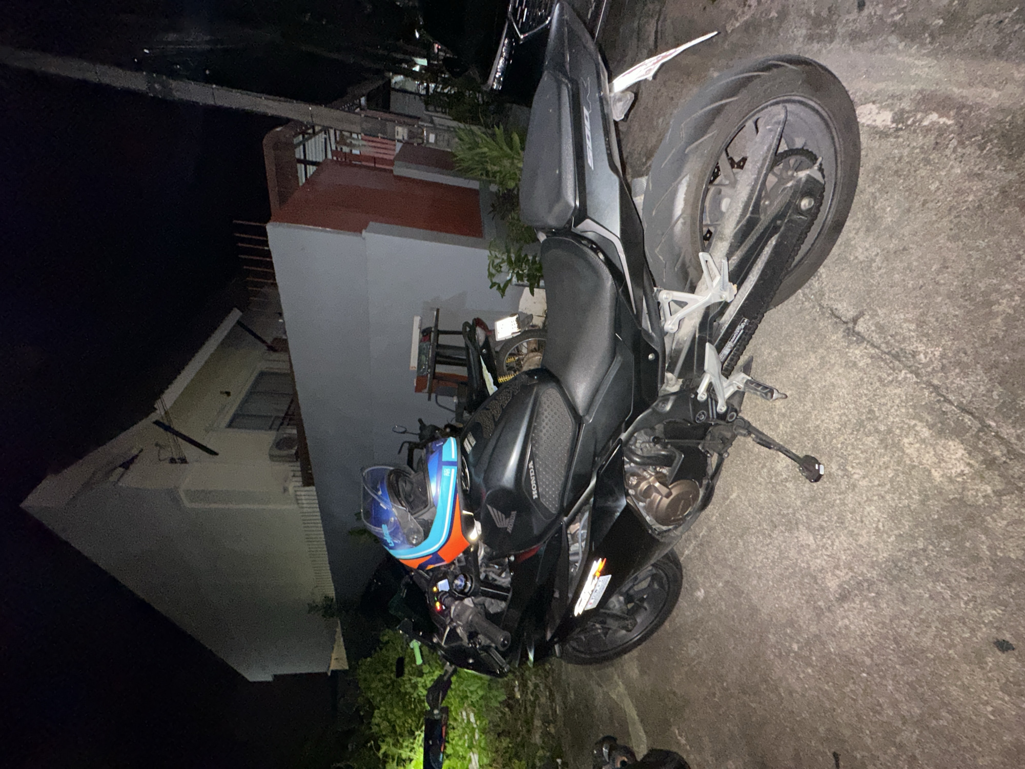 A black Honda motorcycle is parked on a concrete surface at night in Kathu, Thailand. A blue and orange helmet rests on the handlebars of the motorcycle. The motorcycle has a sleek, sporty design with prominent fairings and a visible engine. The scene appears to be a residential area, with a building visible in the background that has red accents and white walls. There is some vegetation to the left of the motorcycle. The lighting suggests it is nighttime, and the overall atmosphere is quiet and still. The image is taken from a slightly elevated angle, looking down at the motorcycle. Some text is visible on the motorcycle, including "HONDA" on the fuel tank.