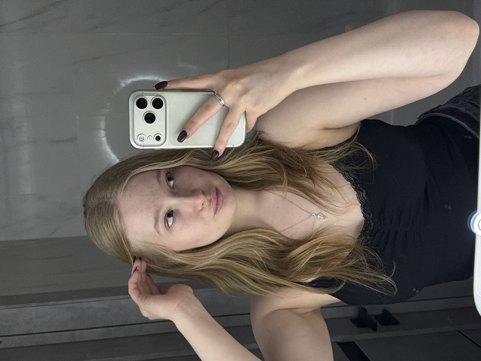 A young woman with long blonde hair and fair skin is captured in a mirror selfie, holding a light-colored smartphone with a prominent camera array in her right hand. Her head is tilted, and she looks directly into the mirror with a neutral expression. Freckles are visible across her nose and cheeks. She is wearing a black top with delicate lace trim along the neckline and a silver-toned necklace with a detailed pendant. A ring is visible on the ring finger of her right hand, and her nails are painted a dark maroon or burgundy color. Her left hand is raised towards her hair, with dark nail polish also visible on those fingers. The background, reflected in the mirror, appears to be an indoor setting with light-colored, possibly tiled or marbled walls. A circular light reflection, possibly from a ring light or overhead fixture, is visible on the right side of the mirror. The image seems to have been taken in Yuzhny, Russia. No visible text is present in the image.