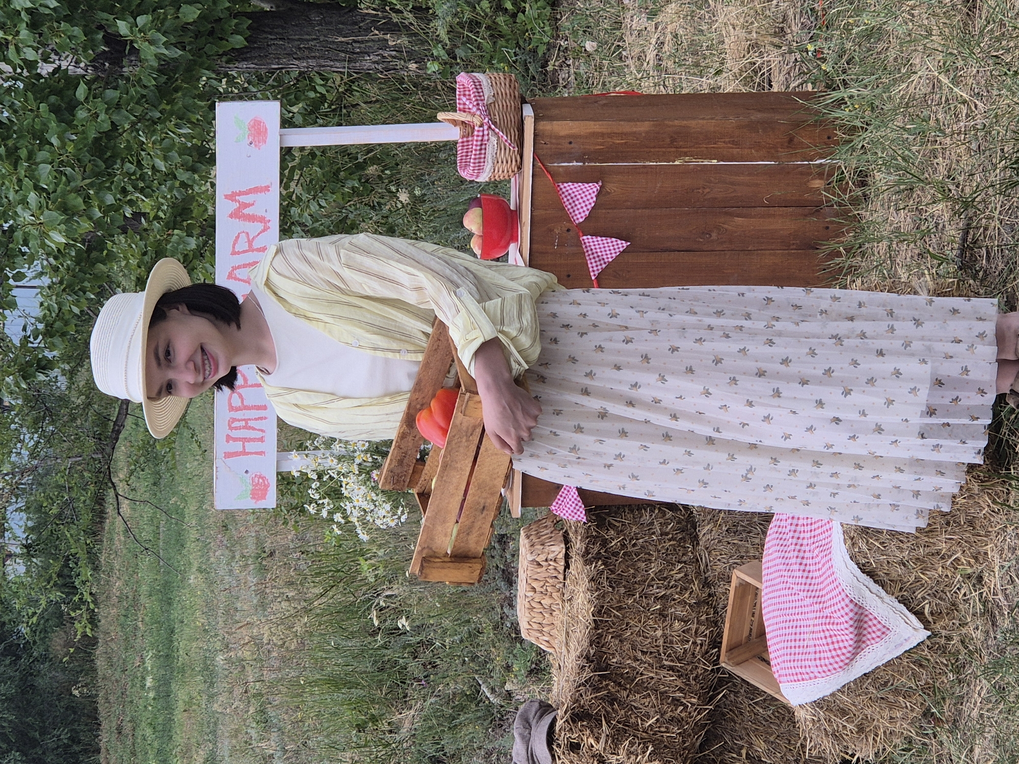 A young woman stands in an outdoor setting, possibly a farm or market stall, holding a wooden crate filled with red bell peppers. She is wearing a white, wide-brimmed hat, a light-colored, long-sleeved blouse with yellow stripes, and a long, flowing white skirt with a subtle floral pattern. Behind her, a rustic wooden stall is decorated with pink and white checkered bunting. On the stall, there is a red bowl containing apples and a woven basket with a pink and white checkered lining. A sign above the stall reads "HAPPY FARM" with a painted strawberry and raspberry. To the left of the woman, several hay bales are stacked, with a straw hat and a checkered fabric draped over them. The background consists of a grassy field with tall dry grass and scattered trees, suggesting a rural or semi-rural environment. The overall scene evokes a rustic, summery agricultural atmosphere. This scene is located in the city of Kokshetau, Kazakhstan.