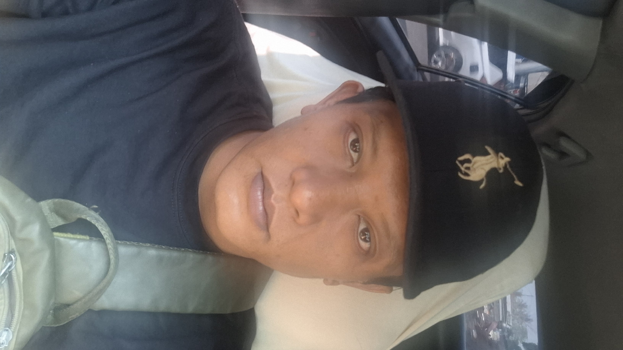 A man is captured in a selfie taken inside a vehicle, possibly a car, in Rancagoong, Indonesia. He is wearing a black baseball cap with a gold polo rider logo and a black t-shirt. A strap of a tan sling bag is visible across his chest. The background shows a blurred view of the outside environment, including a white vehicle and some trees, suggesting a daytime setting. The man's face is the primary focus of the image, with his expression neutral.