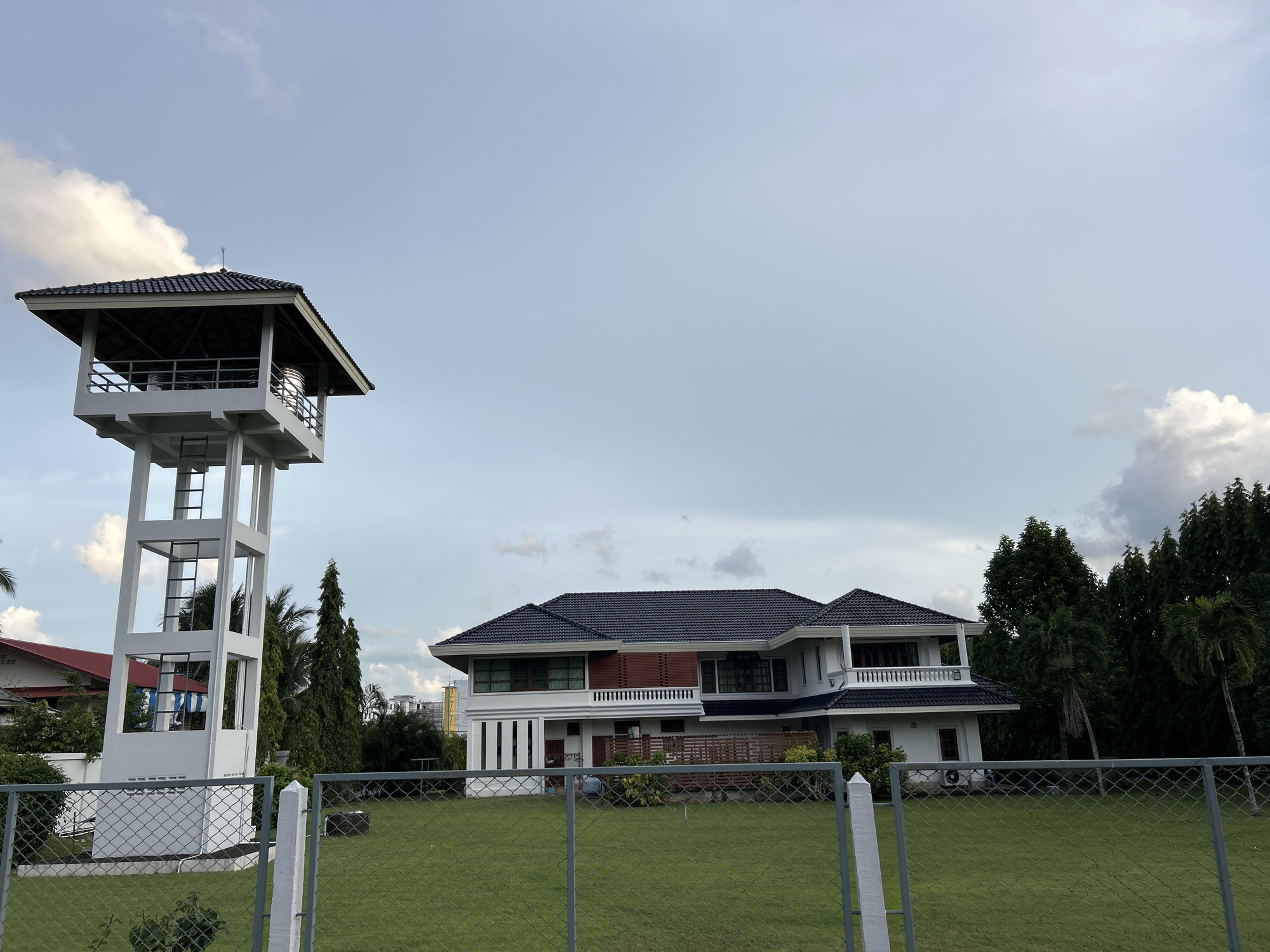 This is a wide shot of a residential property in Kathu, Thailand, under a partly cloudy sky. On the left, a white, multi-story water tower with a dark-tiled roof stands tall. It has an open-air platform with railings at the top, containing two large cylindrical water tanks. A ladder is visible ascending the tower's structure. In the center and right of the image is a large, modern white house with a dark tiled roof. The house has multiple levels, balconies with railings, and large windows. Green grass covers the foreground and the yard of the property. A chain-link fence with white fence posts runs across the front of the yard. Behind the house, a dense line of tall green trees forms a natural boundary. In the distance, other buildings and construction can be seen. The overall scene depicts a well-maintained property with significant infrastructure.