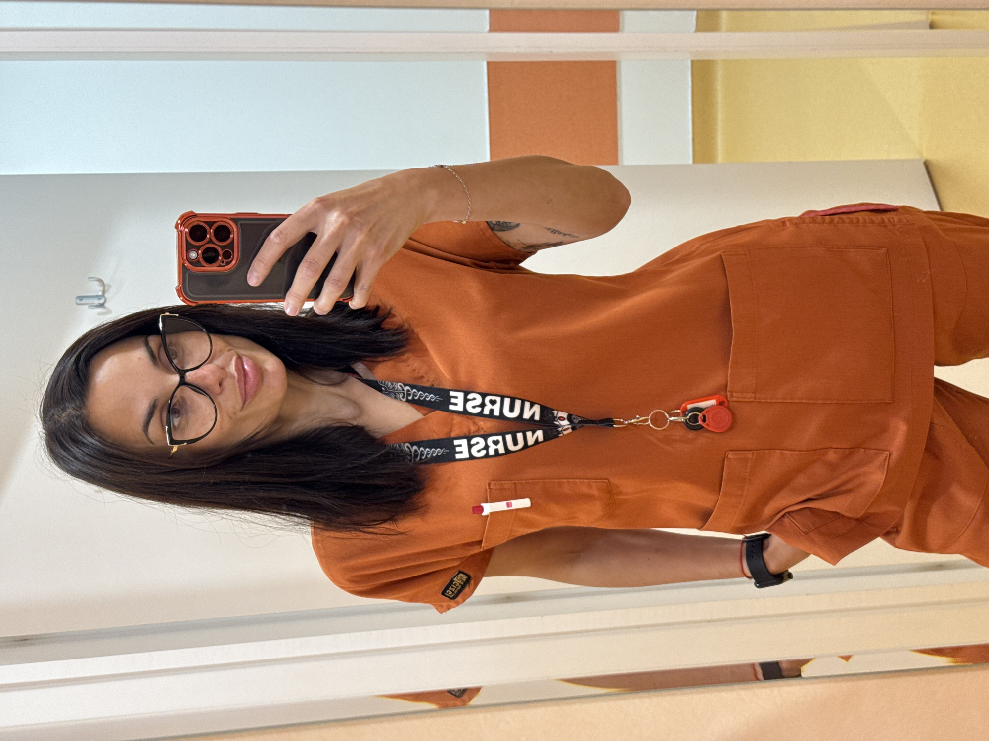 A woman is taking a selfie in a mirror. She is wearing an orange scrub set, glasses, and a lanyard that reads "NURSE" in reverse. She is holding a black and red phone. The background shows a wall with horizontal sections of white, orange, and yellow. The location is presumed to be in a medical facility in Pleven, Bulgaria based on the subject's attire.