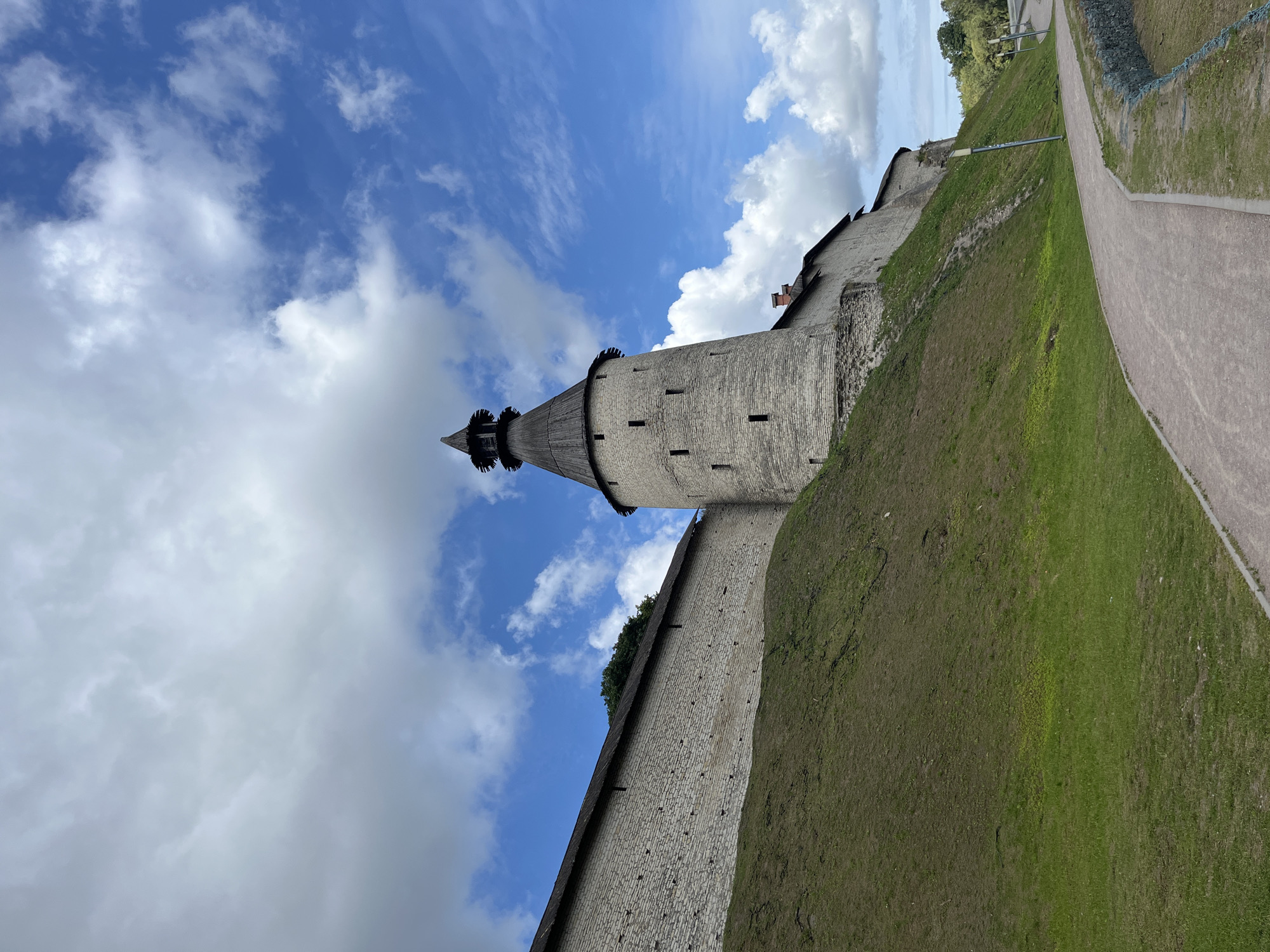 The image depicts a section of the Pskov Kremlin in Pskov, Russia, under a partly cloudy blue sky. A tall, stone watchtower with a wooden, conical roof dominates the right side of the frame, appearing to be part of a larger fortress wall. The wall, constructed from rough-hewn stones, extends to the left, curving upwards along a steep, grassy slope. A paved pathway winds along the bottom of the slope, leading away from the viewer. The environment is verdant, with patches of green grass covering the hillside and a line of trees visible in the distance. No people are present in the image.