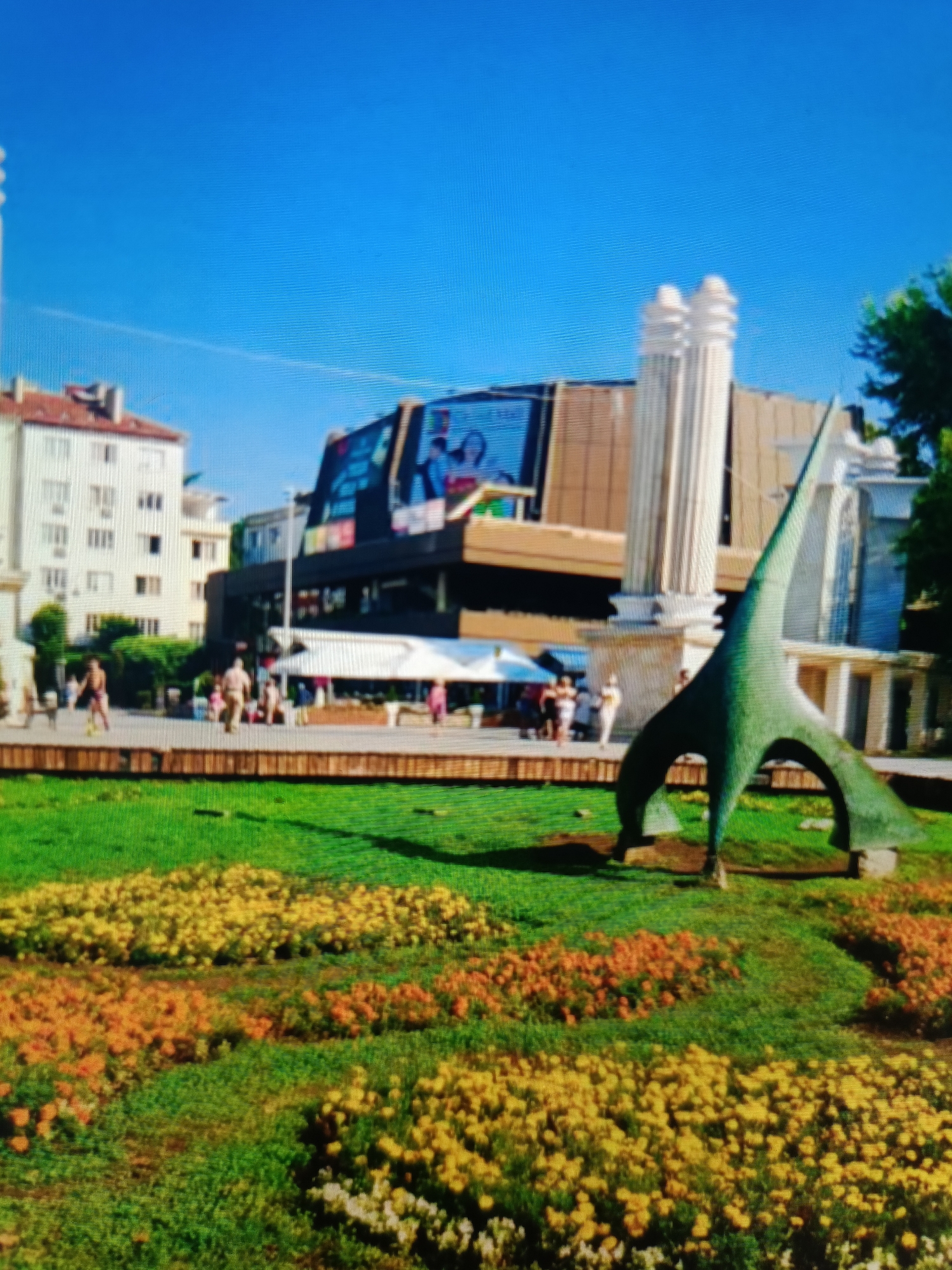 The image depicts a sunny day in Varna, Bulgaria, specifically the area around the Varna Opera House and its adjacent garden. In the foreground, a meticulously maintained garden bed features rows of yellow and orange flowers interspersed with green grass. A striking green abstract sculpture, resembling a stylized bird or creature, is centrally positioned in the garden.
In the midground, people are visible strolling and gathered in an open plaza. A white tent or awning suggests a market or outdoor seating area. Behind the plaza, the facade of the Varna Opera House is prominent, characterized by a modern, angular design with large windows and a screen displaying advertisements or movie posters. To the left of the Opera House, a multi-story residential building with a reddish-tiled roof stands.
Further to the left, two tall, fluted white columns frame the Opera House. The sky above is a clear, vibrant blue, indicating good weather. The overall scene portrays a lively urban environment with a blend of modern architecture, public art, and recreational green space.