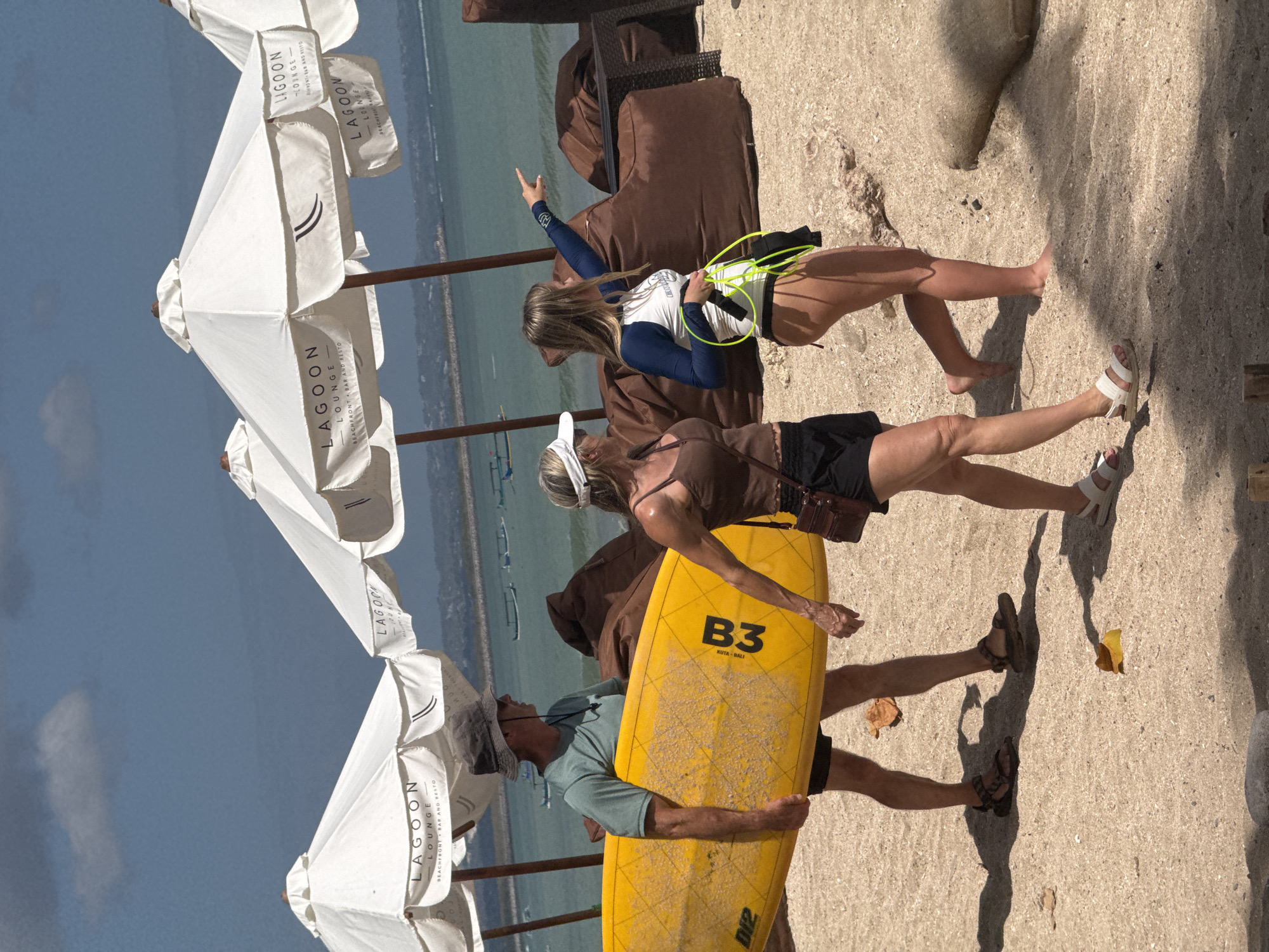 In Kuta, Indonesia, three individuals walk on a sandy beach adjacent to a body of water. A man in a grey bucket hat and teal t-shirt carries a bright yellow surfboard under his right arm. Beside him, a woman in a brown tank top and black shorts walks with a brown crossbody bag. To their right, a young woman wearing a blue and white long-sleeved rash guard and black bikini bottoms points towards the water, holding a coiled green rope. In the background, several white umbrellas with the "Lagoon Lounge" logo are set up, and large brown beanbag chairs are visible. The ocean stretches out under a partly cloudy sky.