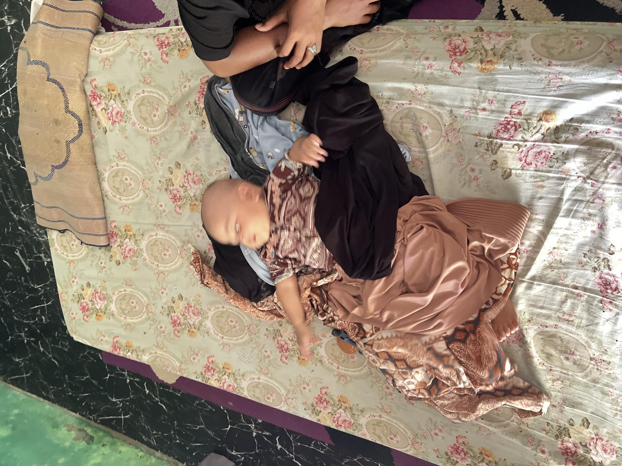 An overhead shot shows a baby lying on a floral-patterned bedspread. The baby is wearing a patterned shirt and dark pants. Scattered around the baby are several articles of clothing, including a brown patterned blanket, a pinkish-brown pleated garment, and some blue patterned fabric. To the right of the baby, an adult's arm and hand are visible, with a ring on the index finger. The background consists of a dark, patterned floor and a section of a purple rug. The scene appears to be indoors, possibly in a bedroom. The image was taken in Lhokseumawe, Indonesia.