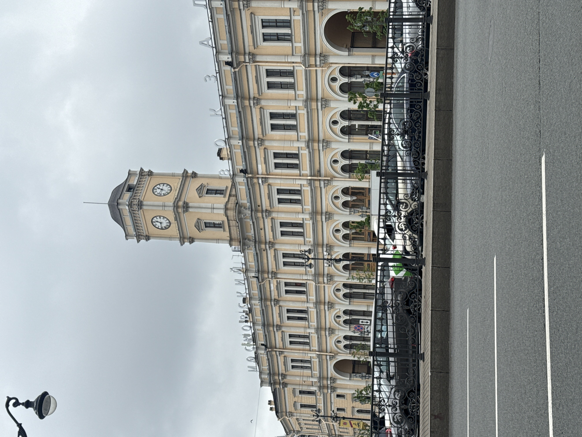 Here is a description of the image:
The image shows the Moskovsky Railway Station in Saint Petersburg, Russia, from a street-level perspective. The building is a prominent architectural feature with a clock tower and a beige facade with several rows of windows and arched openings. The Russian words "МОСКОВСКИЙ" are displayed on the top of the building. A decorative black wrought-iron fence runs along the perimeter. The road is visible in the foreground with white lane markings. Cars and street signs are also visible behind the fence, and a lamppost is partially visible in the lower left corner. The sky is overcast.