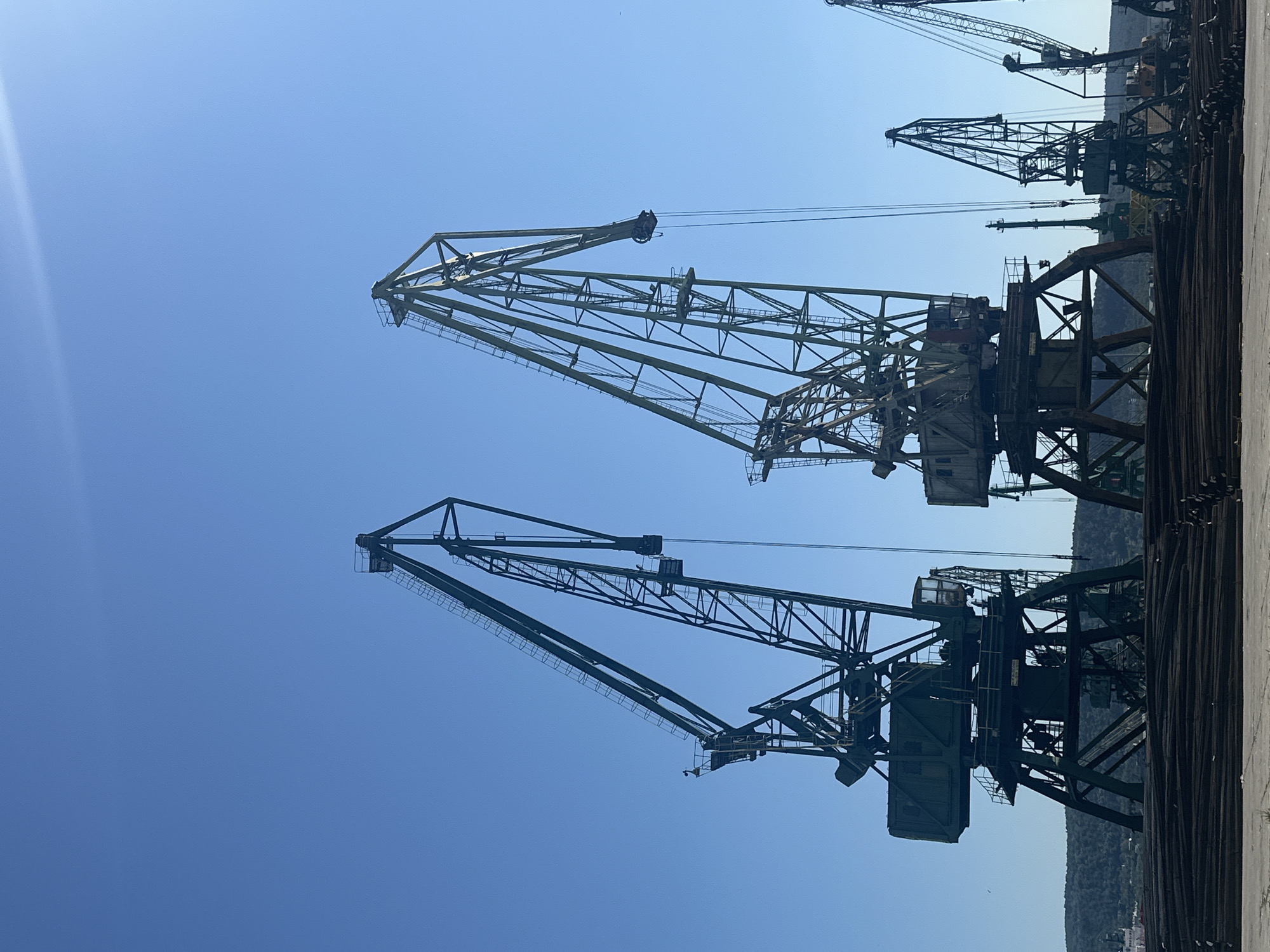 An outdoor, low-angle, medium shot shows two large, dark green industrial cranes silhouetted against a clear, bright blue sky. The cranes are tall and angular, made of a lattice of metal beams, with large, boxy counterweights at their bases. The crane on the left is positioned slightly in front of the crane on the right, and both have long, angled booms that extend upwards and towards the right. The crane on the right has its boom angled more steeply. In the background, a dense line of dark green trees and hills is visible under the horizon. In the foreground, a large pile of dark metal rods is stacked horizontally. Several smaller cranes are visible in the distance, to the right of the two main cranes. The scene appears to be a port or industrial shipping area. There are no people visible in the image.