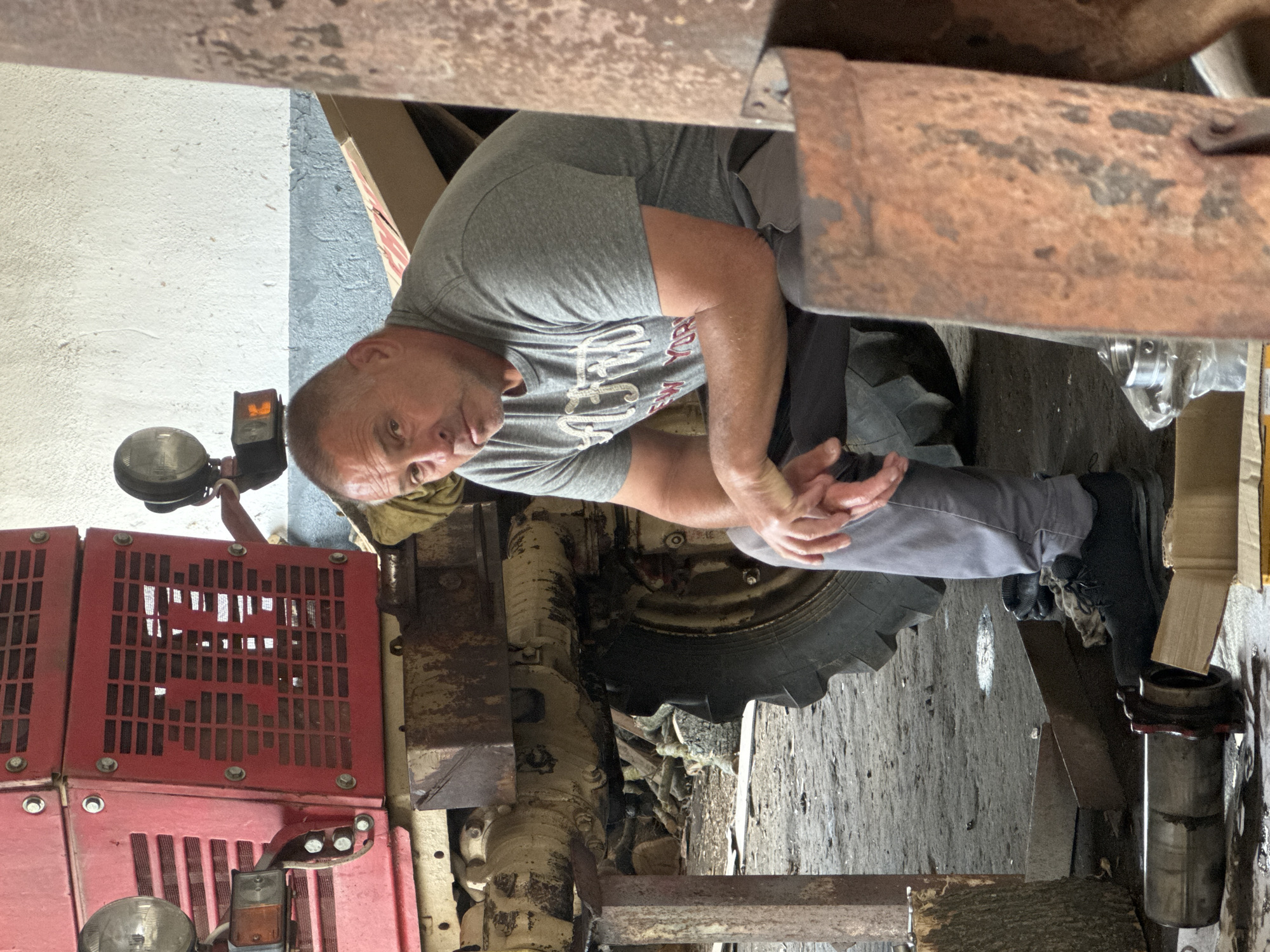 The image, taken in Sarnevo, Bulgaria, shows a middle-aged man seated or crouching next to a large piece of heavy machinery, likely a tractor, in what appears to be a workshop or garage. He is looking directly at the camera with a neutral expression. The man has short hair and is wearing a grey t-shirt with partial text visible, which seems to read "NEW YORK", along with grey pants and dark shoes. His hands are clasped together in front of him.
To his right, a large, dark, lugged tire of the machinery is prominent. The machine's body is a dirty, weathered yellowish-beige, with several rusty metal components, including a substantial rusty beam or pipe above the man's head. On the left side of the frame, bright red metal grates with rectangular cutouts, possibly part of the machine's housing or cooling system, are visible. A round headlight and a square orange signal light are also attached to this side of the machinery.
The background features textured walls, one in a light grey/white and another in a darker grey. The floor is rough, appearing to be concrete or dirt. Cardboard boxes or materials are seen near his feet on the right. The setting suggests an indoor, utilitarian environment, consistent with maintenance or storage of agricultural equipment. The lighting indicates it is daytime.