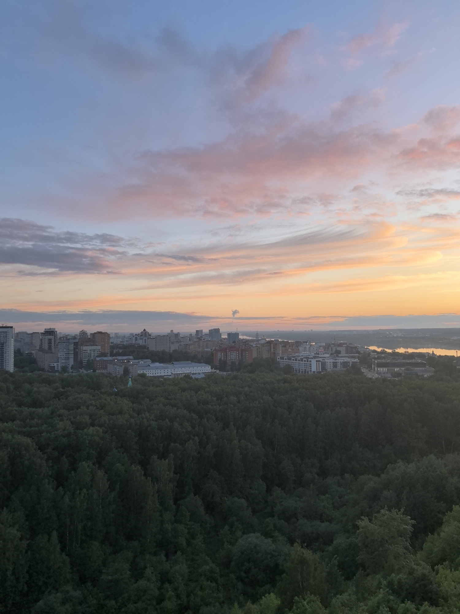 An eye-level outdoor shot depicts a cityscape at dawn or dusk. Predominantly green, dense foliage covers the foreground, suggesting a large wooded area. Behind the trees is a skyline of urban buildings, varying in height and architectural style. Farthest back, a body of water, possibly a river, borders the city. In the sky, gradients of blue transition into streaks of orange and pink clouds near the horizon. This scene is specifically located in Perm, Russia.