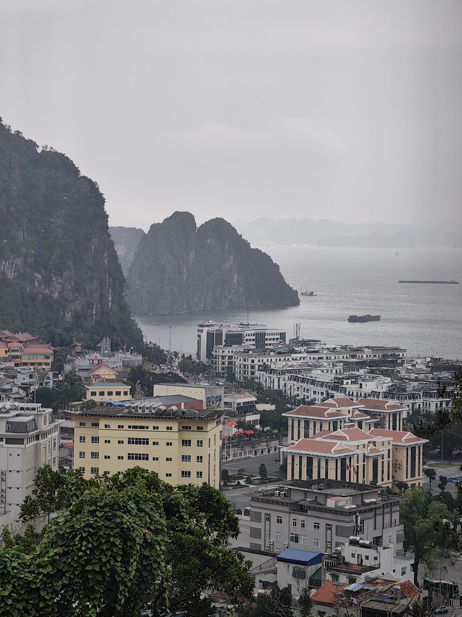 The image captures a general view over Hong Gai Ward, Vietnam, with limestone karsts extending into the sea. In the foreground, a mix of older residential buildings and newer constructions, including large multi-story structures with red-tiled roofs, are visible. Dominating the view are the limestone formations characteristic of the region, shrouded in mist. Several vessels are present in the water, including a barge. The overall atmosphere is overcast and hazy.