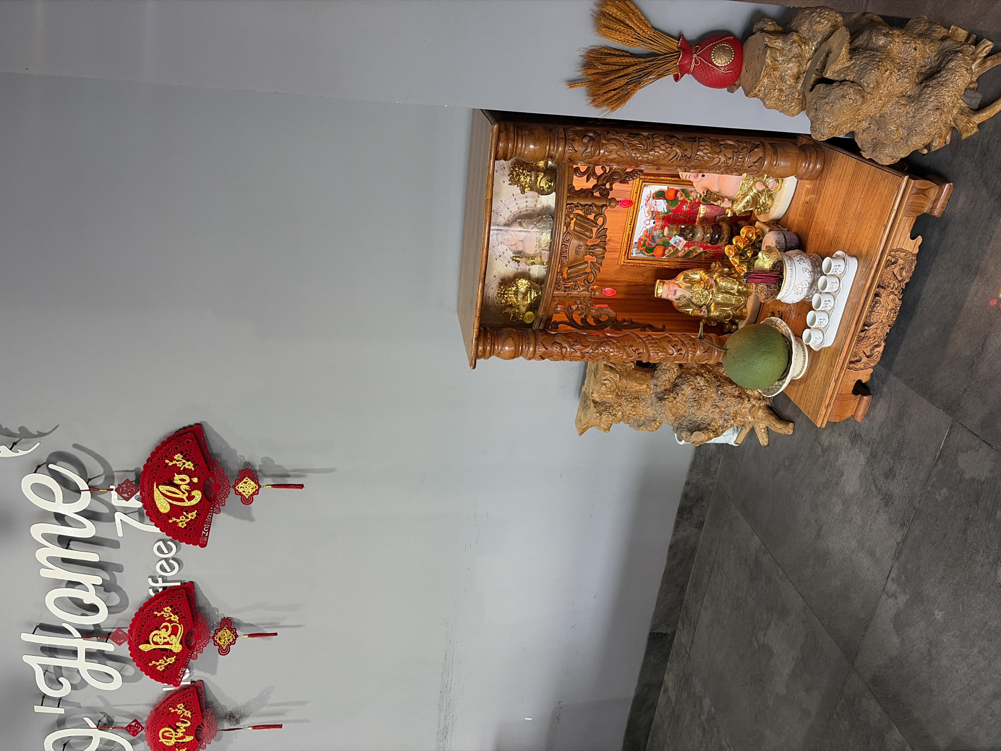 The image depicts an indoor scene, likely a residential or commercial space in Da Nang, Vietnam, characterized by traditional Vietnamese decorative elements. A light gray wall serves as the backdrop for several red, fan-shaped ornaments with gold lettering. Below these, a wooden altar stands prominently, adorned with golden figurines of deities, possibly representing wealth and prosperity. A framed image of a religious scene is placed centrally on the altar. To the left of the altar, a green, round fruit rests on a small white dish, accompanied by a set of five small white teacups arranged on a tray. A decorative wooden sculpture is positioned to the left of the altar. To the right of the altar, a bundle of dried wheat stalks is arranged in a red vase atop another wooden sculpture. The floor is tiled with dark gray square tiles.