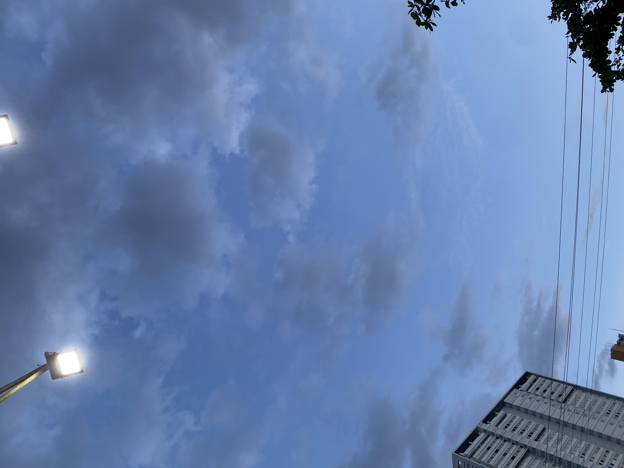 The image depicts a cloudy sky at dusk, with a modern building visible in the lower right corner. A street light illuminates the left side of the frame, casting a bright glow against the darkening sky. Several power lines stretch across the upper right portion of the image, with hints of dark green foliage peeking from the top edge, possibly indicating trees. The sky is a mixture of dark gray clouds and lighter blue, suggesting it might be overcast or that the sun has recently set. The building appears to be an apartment complex or office building, with many windows visible. There are no people or specific activities depicted, creating a sense of stillness and the transition from day to night. The location context provided is Gambut Barat, Indonesia, which might explain the tropical feel of the foliage and the type of building. The overall mood is calm and possibly a bit somber due to the cloudy and dim lighting.