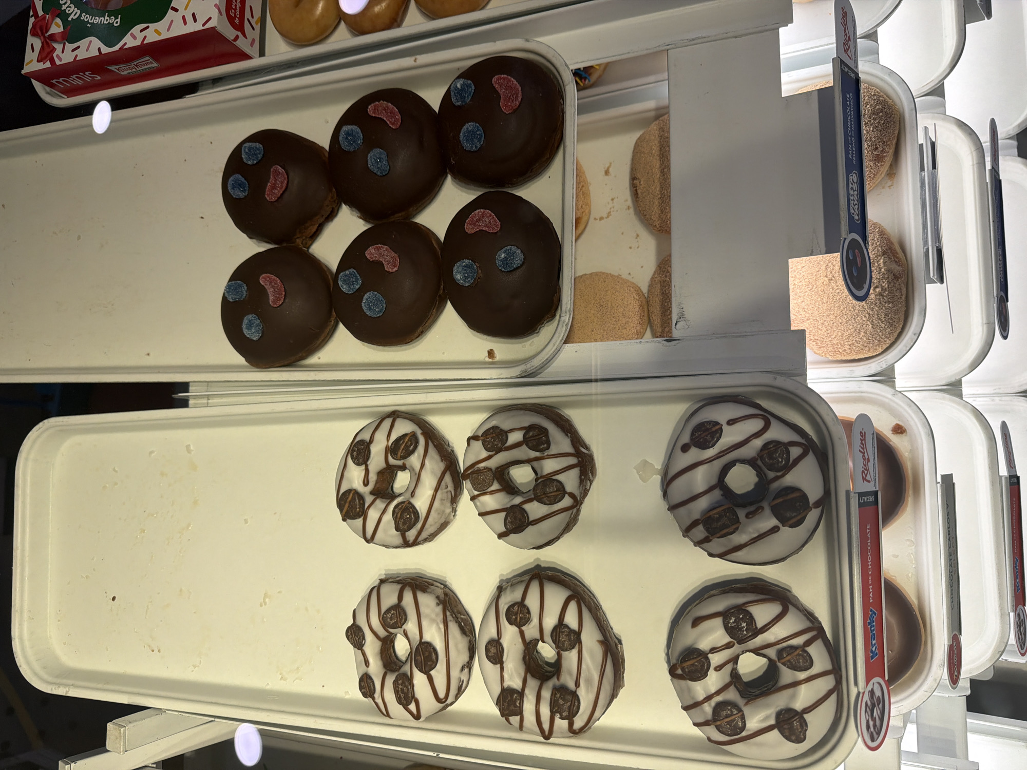 This image, taken in San Bernardino Tlaxcalancingo, Mexico, depicts a commercial display case containing various baked goods. The display is multi-tiered with white, rectangular plastic trays.
The top-left tray is empty, showing faint crumbs and residue on its surface. Directly to its right, the top-right tray holds six dark chocolate-coated, circular pastries, each decorated with two small blue candies for eyes and a wavy red candy for a smiling mouth. To the right of these, two standard glazed, ring-shaped donuts are partially visible, alongside a red and white Krispy Kreme "Minis" box, which is decorated with sprinkles and features text in Spanish.
Below the empty tray, the middle-left tray displays six white-glazed, ring-shaped donuts. These donuts are drizzled with a darker icing and topped with several small, dark brown chocolate pieces. A product label below these donuts identifies them as "Kranky PAN DE CHOCOLATE" (Chocolate Bread) by Ricolino.
Below the smiling chocolate pastries, the middle-right tray contains at least two round, light brown pastries, appearing to be covered in fine crumbs or sugar. A product label for these items, partially obscured, shows a cartoon smiling face and the text "PAN DE CHOCOLATE RELLENO DE CARAMELO" (Chocolate Bread filled with Caramel). Further down, additional obscured trays and product labels, including one for "CHOCOLATE CARLOS V" and another "Kranky PAN DE CHOCOLATE," are visible.
The scene is illuminated by bright, likely overhead, artificial light, casting minimal shadows and highlighting the texture of the baked goods and the clean surfaces of the display. No individuals are present in the image.