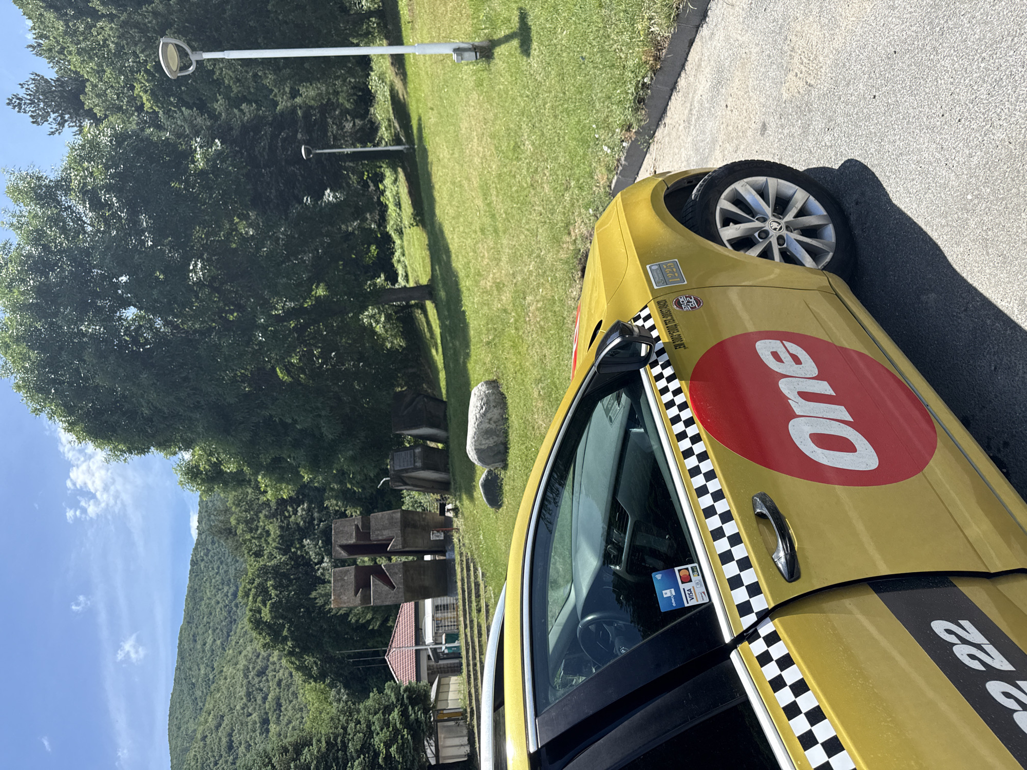 The image displays an outdoor scene featuring a yellow taxi cab parked on an asphalt surface. The taxi, possibly a Skoda given the wheel design, has a prominent red circle with "One" in white letters on its side and a black and white checkered stripe running along its length. Visible text includes payment card logos such as "VISA," "Mastercard," "Postbank," and a contactless symbol on a sticker near the driver's window. The number "22" is partially visible on the rear side of the car.
To the left of the car, a lush green grassy area is bordered by several large, leafy trees. Two modern streetlights stand within the grass. Further back, an abstract sculpture made of dark, block-like structures and a large grey rock are positioned on the lawn. A residential building with a red-tiled roof is partially visible behind these elements. In the distant background, a sprawling forested mountain range rises under a clear blue sky dotted with a few white clouds.
The strong sunlight and distinct shadows suggest it is midday or early afternoon on a clear, sunny day. No people or activity are visible, creating a serene atmosphere. The location cues, including the specific taxi branding and the natural landscape, place the scene in Vetren Dol, Bulgaria.