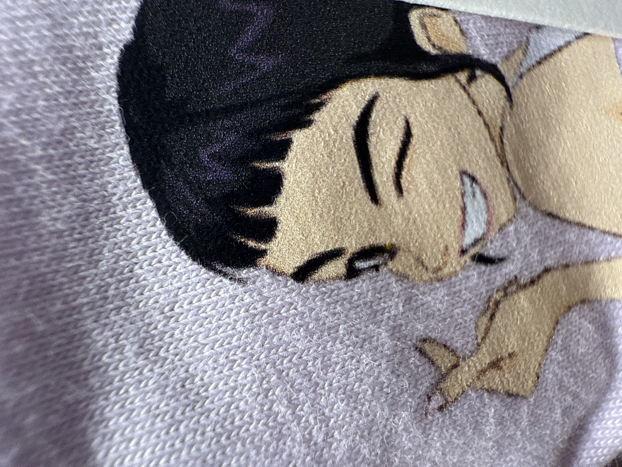 The image is a close-up photograph of a fabric material, likely a blanket or clothing item, featuring a printed cartoon character. The character is depicted in an anime or animated style, showing a head with short, dark black hair and light-toned skin. The character's face is seen in profile or a three-quarter view, with subtle dark lines suggesting closed or resting eyes and a small white detail at the mouth area, possibly a tooth. Part of a hand and arm, rendered in the same light skin tone, are visible in the lower right portion of the image.
The background fabric is a soft, light lavender or pale purple, exhibiting a clear, tight knit or woven texture with visible loops and fibers. The printed elements of the character appear slightly integrated into the fabric's texture, giving the illustration a soft-focus or slightly fuzzy edge rather than a sharp, glossy finish. The overall scene is abstract, focusing entirely on the textile and its printed design, with no discernable external setting or background. There is no visible text in the image. The lighting appears soft, highlighting the texture of the fabric and the details of the character's illustration.