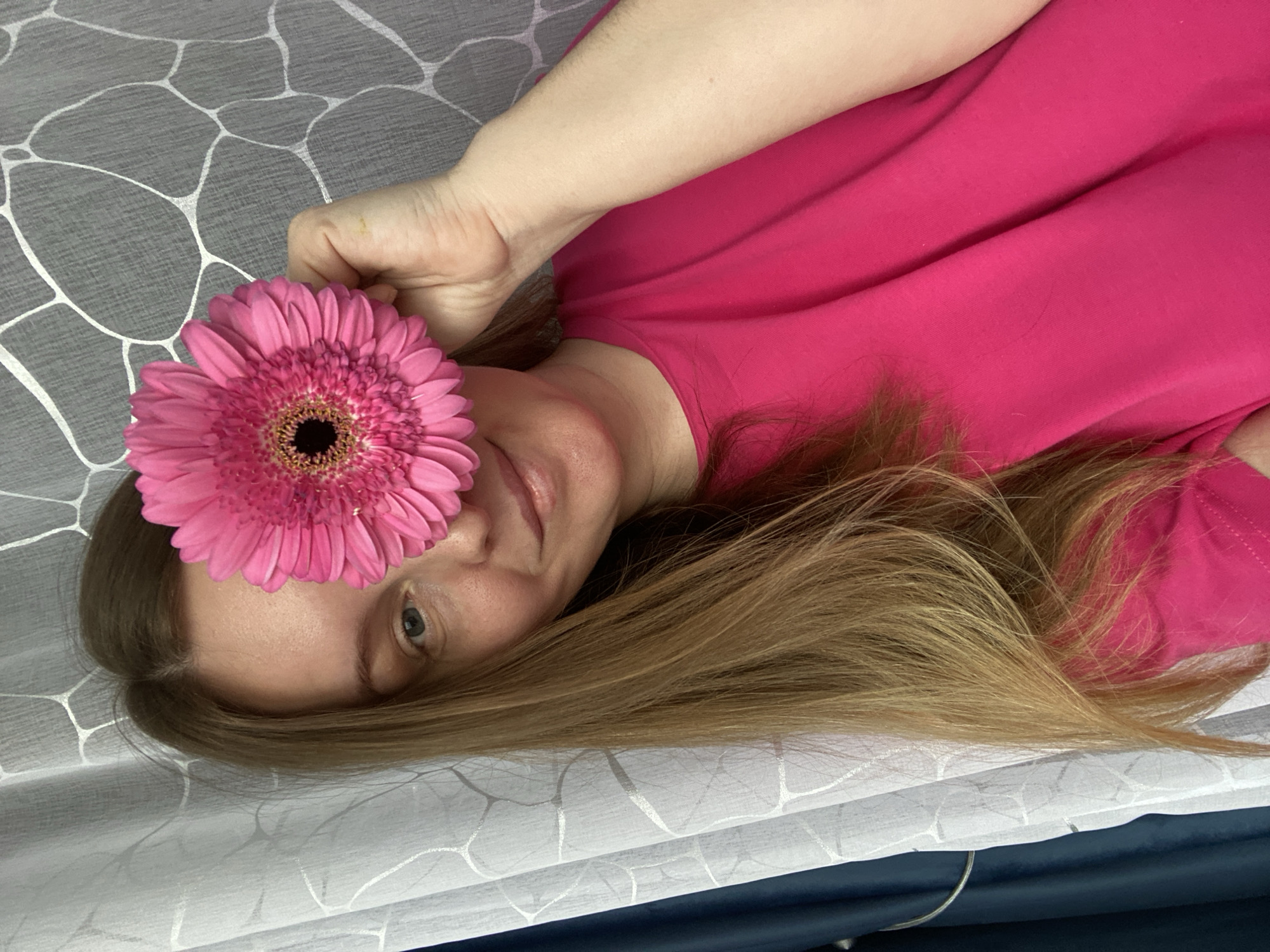 A woman with long blonde hair lies sideways on a patterned gray and white surface, possibly a bed or sofa. She wears a bright pink top and is holding a vibrant pink gerbera daisy in front of her left eye. Her expression is slightly smiling. The location is not specified and it is not possible to determine from the image that it is in Noviy, Russia.