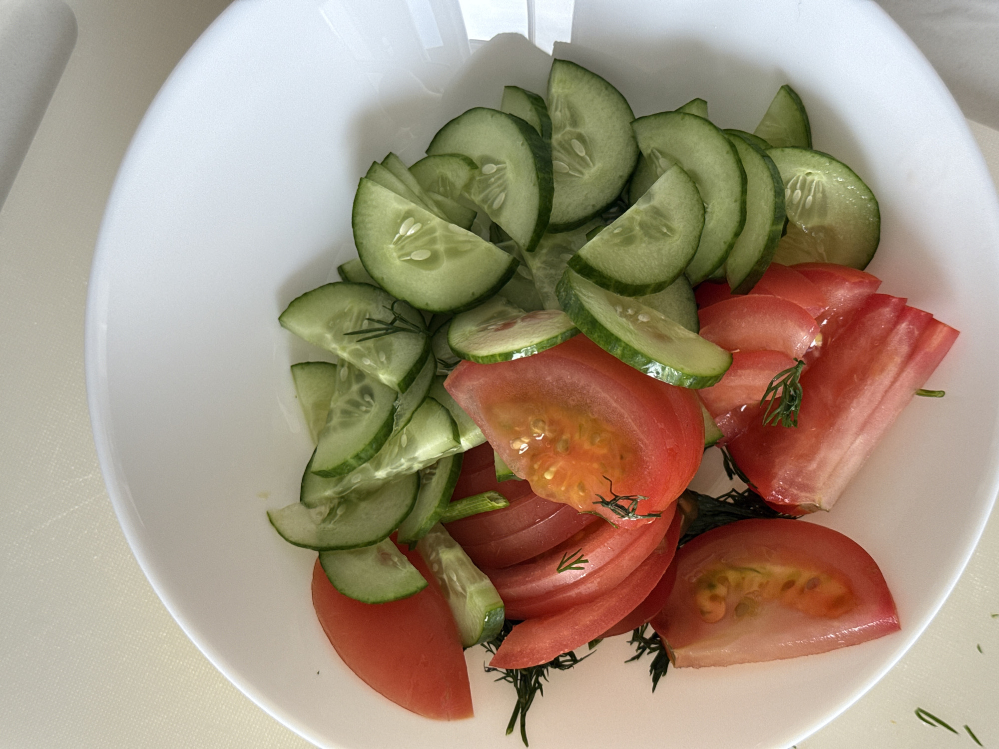 In a white bowl, a salad is arranged. Sliced cucumbers, with their green skins and seedy centers, are mixed with wedges of red tomatoes. Sprigs of dill are interspersed among the vegetables. The bowl sits on a light-colored surface. The location is unspecified.